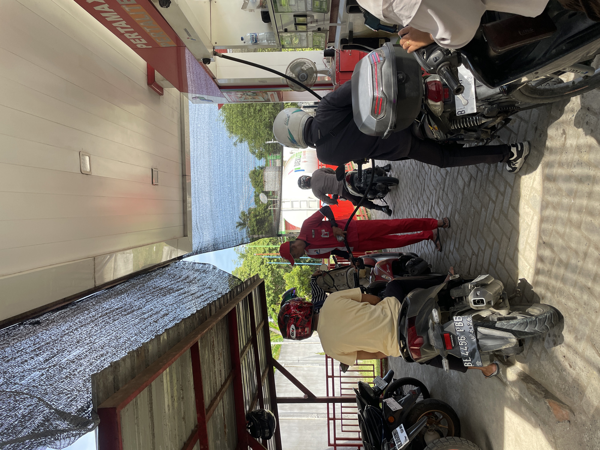 The image depicts a scene at a gas station in Glee Gogo, Indonesia. Several people on motorcycles are waiting to refuel. A gas station attendant, wearing a red uniform and cap, is seen holding a gas nozzle and attending to a motorcycle. The background shows a white building with "PERTAMAX" signage, indicating it is a Pertamina gas station. Trees and a blue sky are visible in the distance. The ground is paved with light-colored bricks.