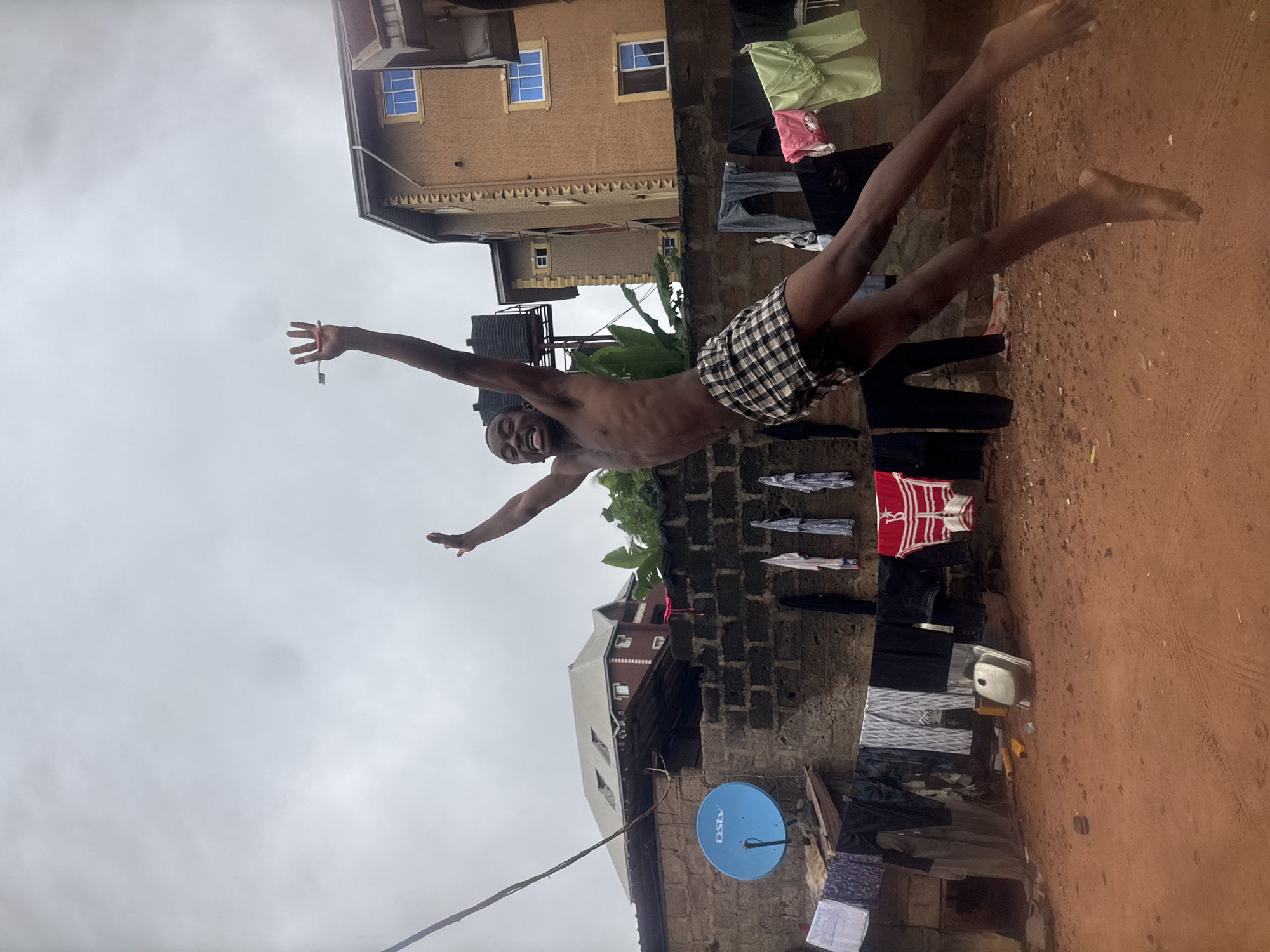 In Nkpor, Nigeria, a shirtless young Black male, wearing plaid shorts, is captured mid-air in a jump, his arms outstretched and mouth open in a joyful expression. He is holding a toothbrush in his right hand, pointed upwards. The scene takes place in a cluttered outdoor area with a red dirt ground. Clothes are hung on a line in the background, and a blue satellite dish is visible on a brick wall. A multi-story building stands behind the clothesline, and another building with a grey roof is seen further in the distance. The sky is overcast and grey.