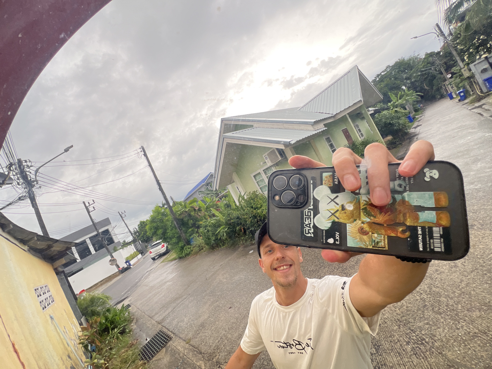 In this media file, a smiling man wearing a white t-shirt and a black baseball cap is taking a selfie outdoors. He is holding up a smartphone with a patterned case featuring a stylized character. The man appears to be on a street, with a light green house and lush greenery visible in the background. The sky is overcast with dramatic clouds, suggesting it might be a cloudy or rainy day. Electrical wires and poles are also present, indicating an urban or suburban setting. The ground appears wet, possibly from recent rain. The overall impression is a candid moment captured during an outing, with a sense of casualness and good spirits. Visible text on the phone case includes stylized lettering and what appears to be a barcode. The location context provided is Choeng Thale, Thailand.