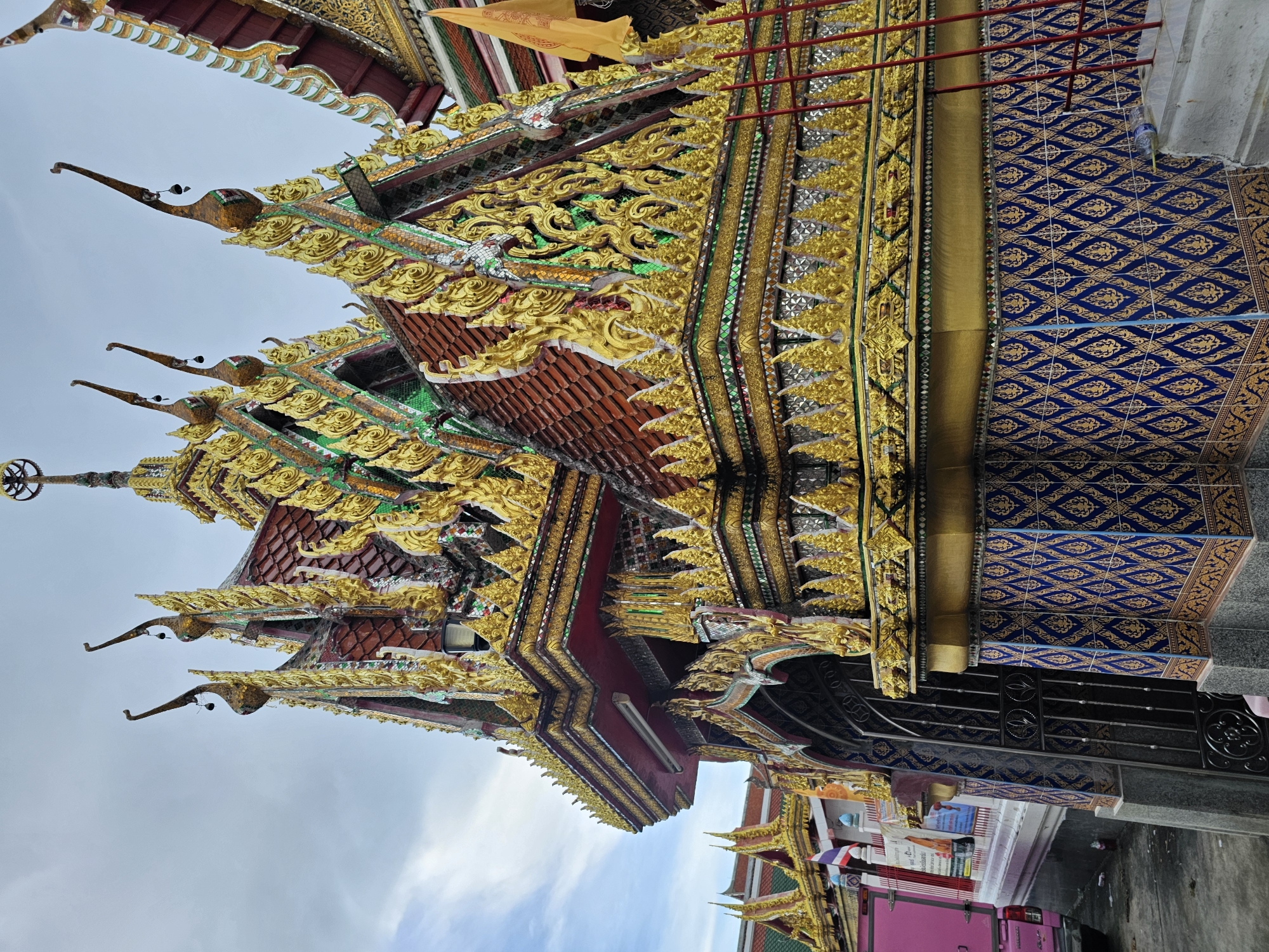 The image depicts the ornate exterior of a Buddhist temple in Bangkok, Thailand. The temple's roof is adorned with intricate golden carvings and red tiles, featuring decorative finials resembling mythical birds with hanging bells. The lower facade is decorated with blue tiles featuring a repeating gold diamond pattern. To the left, a pink truck and a section of a white fence with Thai flags are visible. A black metal gate is present to the left of the main temple structure. A yellow flag is visible on the right side of the image. The sky is overcast with a few patches of blue.