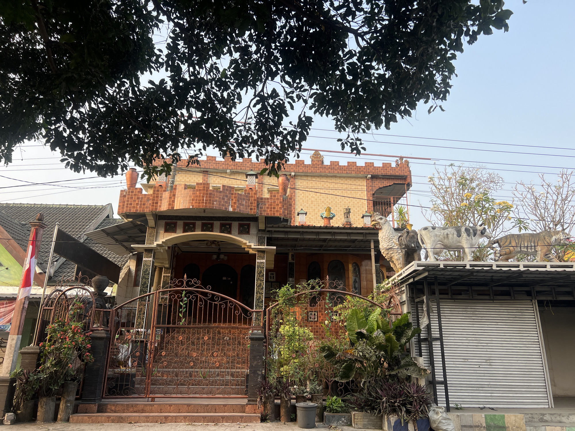 The image depicts a residential property in Tlogoadi, Indonesia, characterized by its unique architectural style and decorative elements. The facade of the house is constructed with red brick and features ornate details, including what appears to be a crenellated parapet reminiscent of a castle. Several decorative elements are visible, including statues of animals such as a chicken, an elephant, a cow, and a tiger, positioned on a metal structure above a roll-up door. The entrance to the property is secured by a decorative metal gate adorned with floral motifs, and the grounds are landscaped with various plants and potted flowers. An Indonesian flag is displayed on a pole near the entrance. The overall scene suggests a personalized and somewhat eclectic approach to home design, with a blend of traditional and whimsical elements. The sky is clear, indicating a daytime setting.