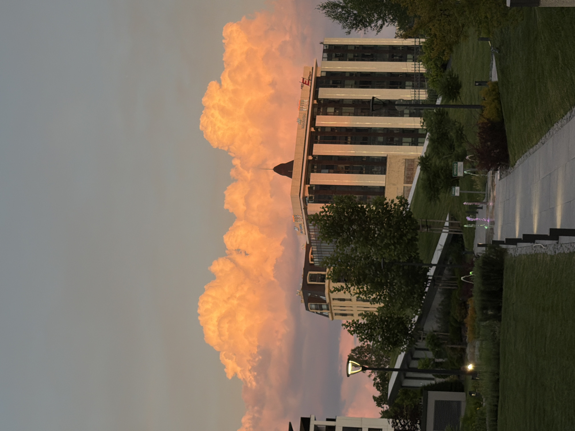 The photograph captures a scene in Sofia, Bulgaria, during what appears to be sunset. Dominating the sky are large, luminous clouds tinged with orange and pink hues, indicative of the setting sun. In the middle ground, a modern office building with a beige facade and vertical white accents stands prominently. The building features numerous windows and a dark, pyramidal structure on its roof. To the left of this building, another structure, partially visible, is a modern building with a white exterior and dark windows.
The foreground depicts a well-maintained park area with lush green grass. A paved walkway extends into the scene, bordered by low plantings and decorative grasses. Several lampposts are visible throughout the park, one with a light illuminating the path. In the distance, within the park, there are hints of a fountain with colored water jets. The surrounding vegetation consists of mature trees, some of which appear to be deciduous. The overall impression is one of an urban park setting with contemporary architecture set against a dramatic sunset sky. There are no people visible in the image.