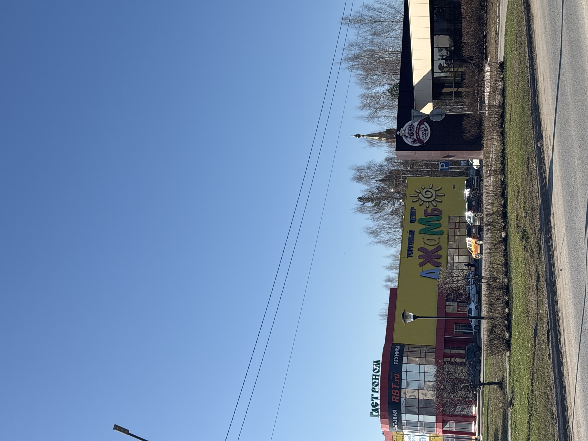The image displays a street scene on a clear, bright day, rotated 90 degrees clockwise. When viewed upright, the upper portion is dominated by a vast, cloudless blue sky, crossed by several utility lines.
Along the right side of the image, there is a paved road flanked by a strip of green grass and patches of bare soil, indicating early spring or late winter. Several buildings line the street.
From left to right (as if viewing upright):
* A multi-story building featuring red and yellow facades. Its signs prominently display "Гастроном" (Gastronom), "RBT.ru", and "БЫТОВАЯ ТЕХНИКА" (Home Appliances), suggesting a grocery store and electronics retailer. A black streetlamp stands in front of this building.
* Adjacent to it, a large yellow commercial building features a sign with a sun logo and the text "ТОРГОВЫЙ ЦЕНТР АЖОМ" (Shopping Center Azhom).
* Further along, a dark-colored building has a circular sign that reads "Лавка cafe" (Shop cafe), with additional, less legible text within the logo, likely "от..." and "Кафе". A blue parking sign ('P') is visible near this building.
* In the background, behind the buildings and bare trees, the ornate steeple of what appears to be a church or similar structure is visible, poking above the horizon.
Bare-branched trees and bushes line the street and are scattered between and behind the buildings, reinforcing the late winter or early spring setting. Several cars are parked in front of the commercial establishments. The overall impression is a sunny, commercial area in Chaikovsky, Russia.
