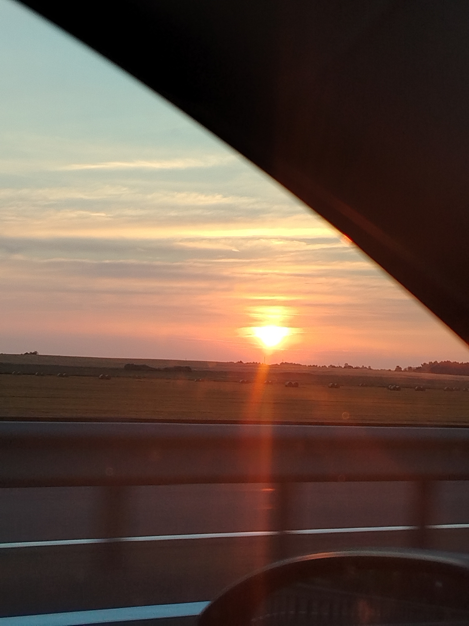 A view through a vehicle window shows a sunset over a rural landscape. The sky is a gradient of pale blue, pink, and orange, with thin, wispy clouds. The sun, a bright orange orb, is near the horizon. In the foreground, a dark, curved object, likely part of the vehicle's interior, frames the scene. Below the sky, a field of dry grass with scattered round hay bales stretches to the horizon. The field appears to be agricultural land. A dark, horizontal line, possibly a road or barrier, runs across the lower part of the image. The lighting suggests it is late afternoon or early evening.