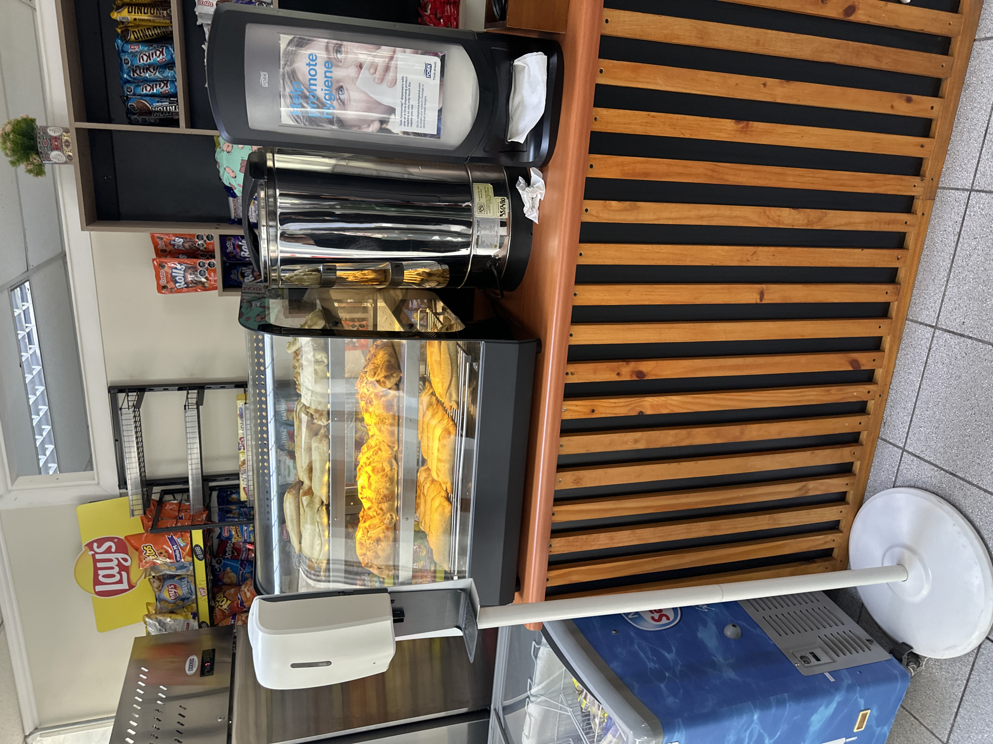 The image shows a commercial food display and preparation area. In a glass-fronted heated display case are rows of baked goods. Behind the display are snacks and drinks. A hand sanitizer dispenser is attached to the front of the display. To the right, a wooden slatted surface is visible, possibly part of the counter or display structure. A napkin dispenser is on the countertop. The location is possibly in Arica, Chile.