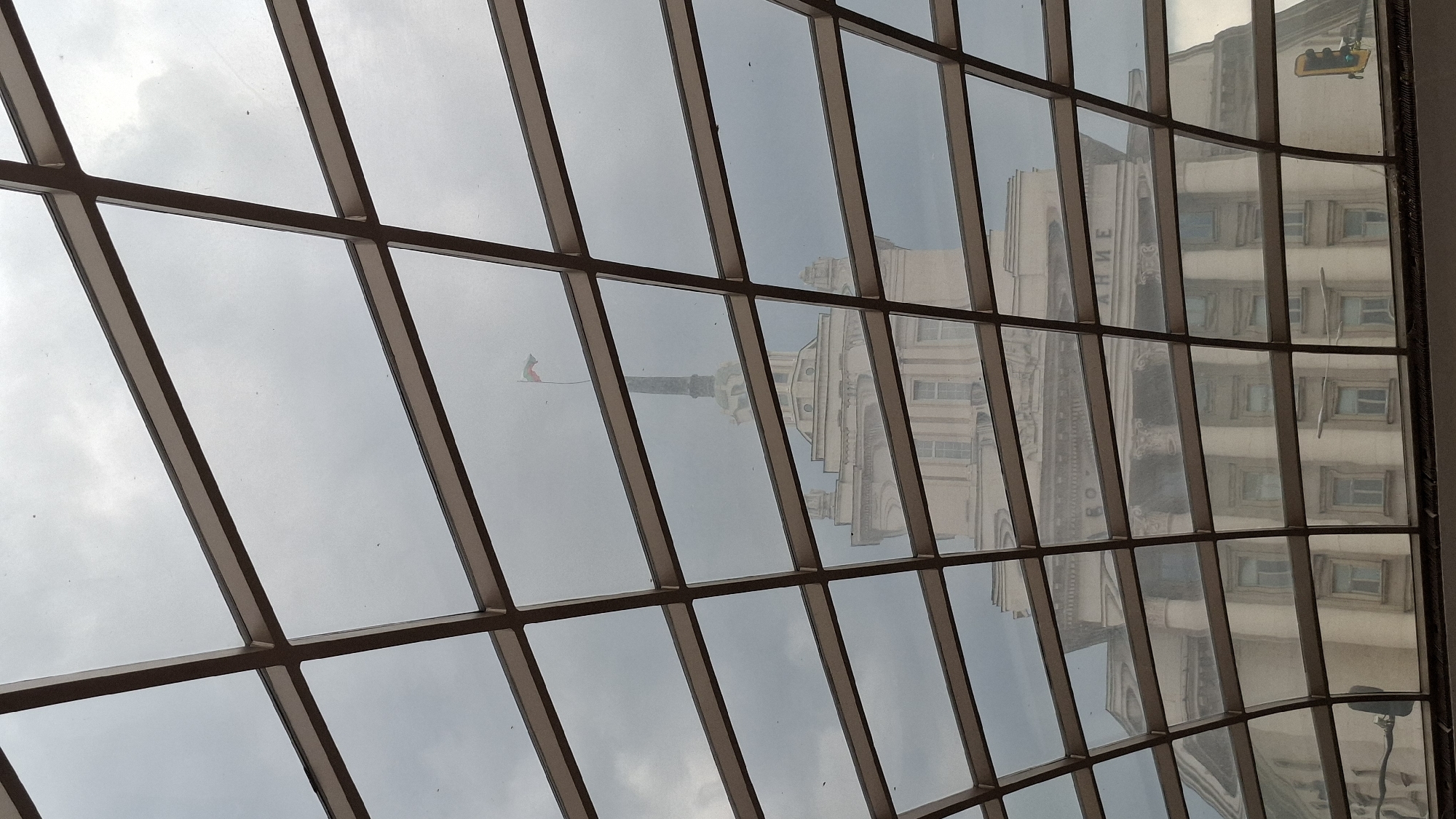 The image depicts an interior view of a glass-paneled ceiling or dome, likely inside a building in Sofia, Bulgaria. The reflection of the exterior environment is visible through the glass panels. This reflection includes the architectural features of the "Largo", specifically the former Party House, with its distinctive columns and facade. There's also a reflection of a flag atop a structure. The metallic framework supports the glass panes. A yellow, wheeled maintenance mechanism is seen near the right edge of the image. The image is taken from an oblique angle.