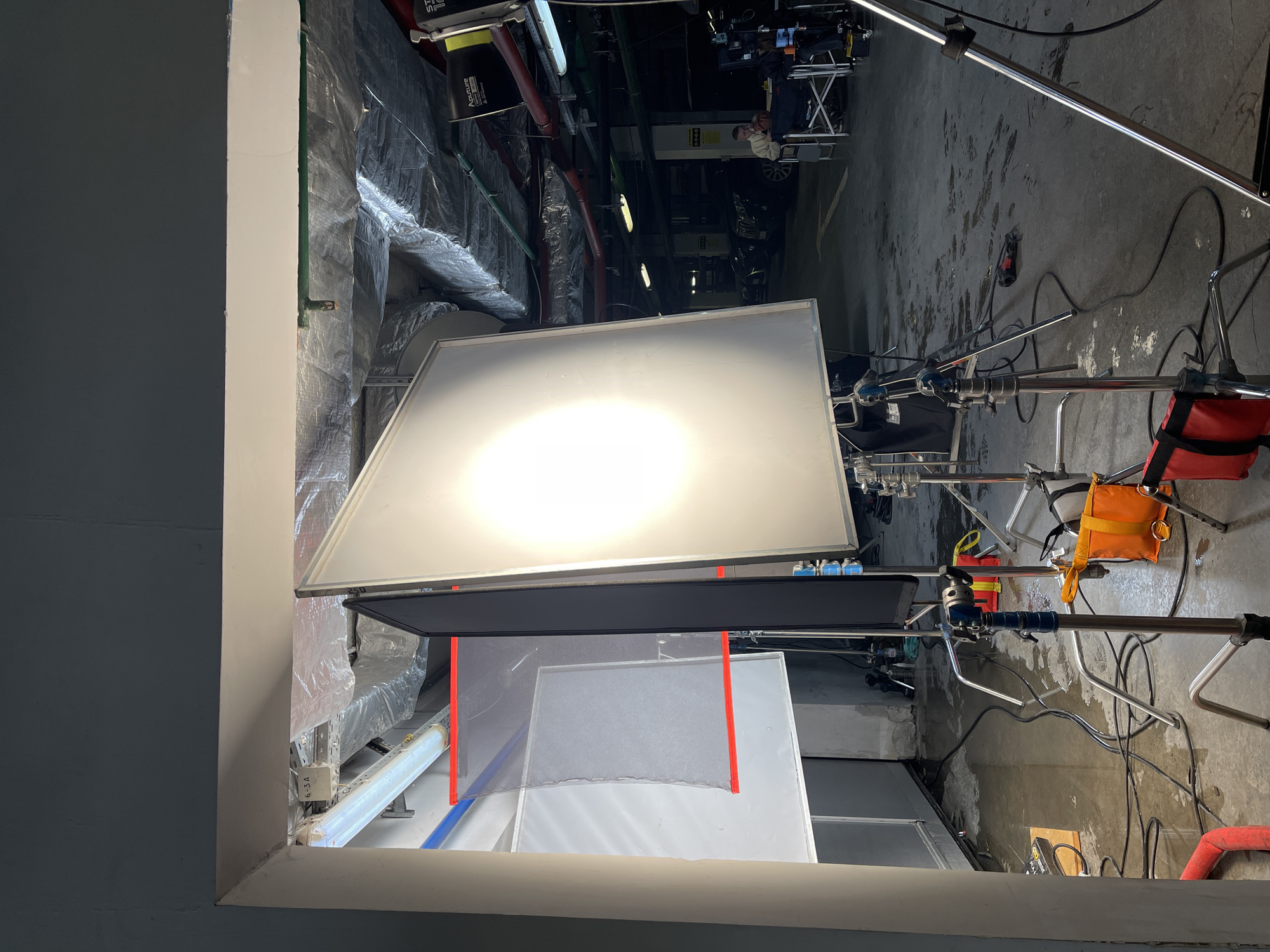 The image depicts a film set with lighting equipment, including reflectors and diffusion screens, in what appears to be a utility or service area. The presence of industrial piping, ductwork, and concrete surfaces suggests a basement or storage space. There is no indication of location. No people are directly visible.