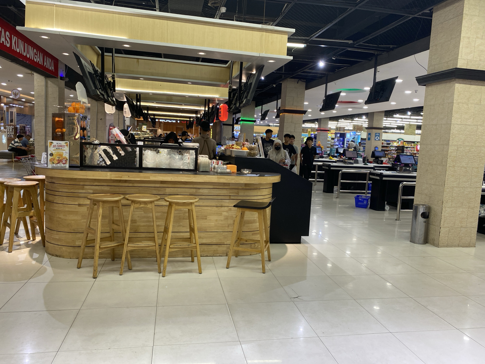 The image captures an indoor scene, likely a food court or a service counter area within a commercial establishment in Banda Aceh, Indonesia. In the foreground, a wooden counter with a polished surface is prominent, featuring several wooden bar stools in front of it. Behind the counter, various food and drink preparation items are visible, including what appear to be juice dispensers with oranges, containers of ice, and other service-related equipment.
Several individuals are interacting at the counter. One person, wearing a brown apron, appears to be working behind the counter. Other individuals, customers, are standing or seated at the counter, suggesting ongoing transactions or service. The overall environment is brightly lit with artificial lighting from above, illuminating the tiled floor and the structure of the establishment.
In the background, more of the commercial space is visible, including rows of shelves stocked with merchandise, suggesting this is part of a larger retail store or a mall. There are also checkout counters with electronic displays. The architectural style features light-colored tiled pillars and a suspended ceiling with exposed beams and lighting fixtures. The presence of a red lantern-like decoration hanging from the ceiling adds a touch of cultural or thematic element to the decor. The scene conveys a busy, functional commercial atmosphere.