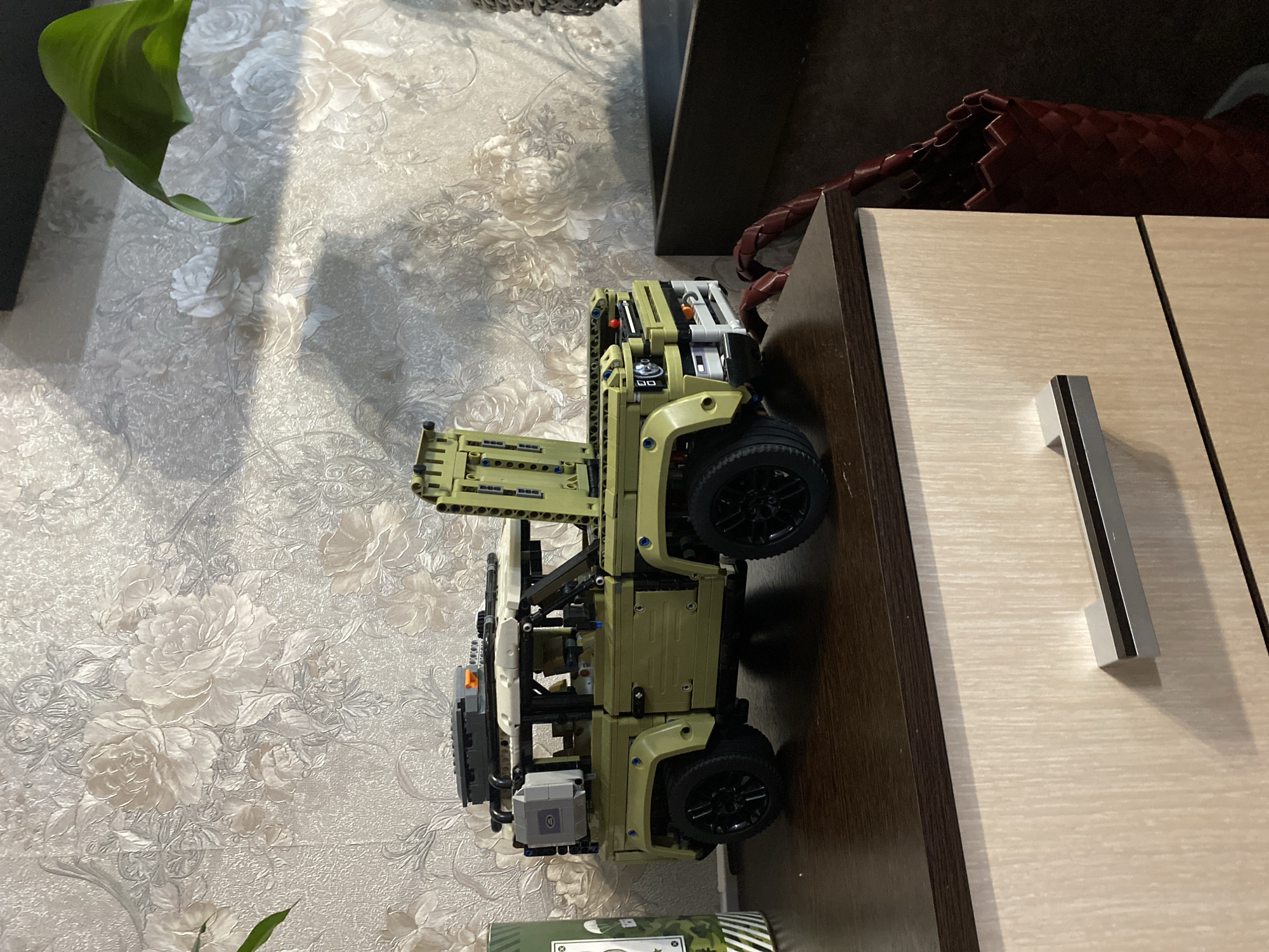 A detailed, forensic, matter-of-fact description of the image:
The image shows a green Lego Technic Land Rover Defender model positioned on the dark brown top of a light-colored wooden dresser with a brushed finish. The dresser has a silver horizontal drawer pull on the visible drawer. The background features a textured wallpaper with a floral pattern in shades of cream and gray. To the left of the model, a green cylindrical object with white stripes and graphics is partially visible. To the right, a dark brown recessed area is present, with a red woven bag inside it. A large green leaf from a houseplant is visible in the upper right portion of the image, casting a shadow on the wallpaper. The lighting suggests an indoor setting with natural light coming from the left. The scene appears to be a still life arrangement on furniture, potentially in a residential environment within the city of Berezovaya Roscha, Russia. There are no people or animals present in the image.