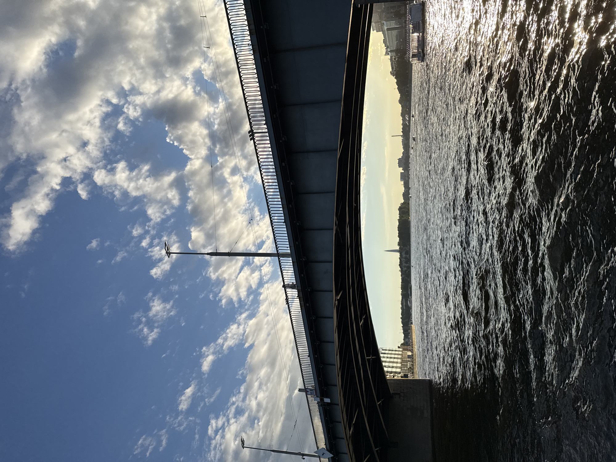 The image captures a view of the Trinity Bridge in Saint Petersburg, Russia, from a boat on the Neva River. The bridge, a prominent landmark, stretches across the frame with its distinctive grey structure and ornate railings. A person is visible walking on the bridge's pedestrian walkway, silhouetted against the bright sky. The river below is choppy, with sunlight glinting off the dark water. In the distance, the city skyline is visible, featuring buildings and the spire of the Peter and Paul Fortress. The sky is a brilliant blue, dotted with fluffy white clouds, suggesting fair weather. The overall scene evokes a sense of urban beauty and the grandeur of Saint Petersburg.