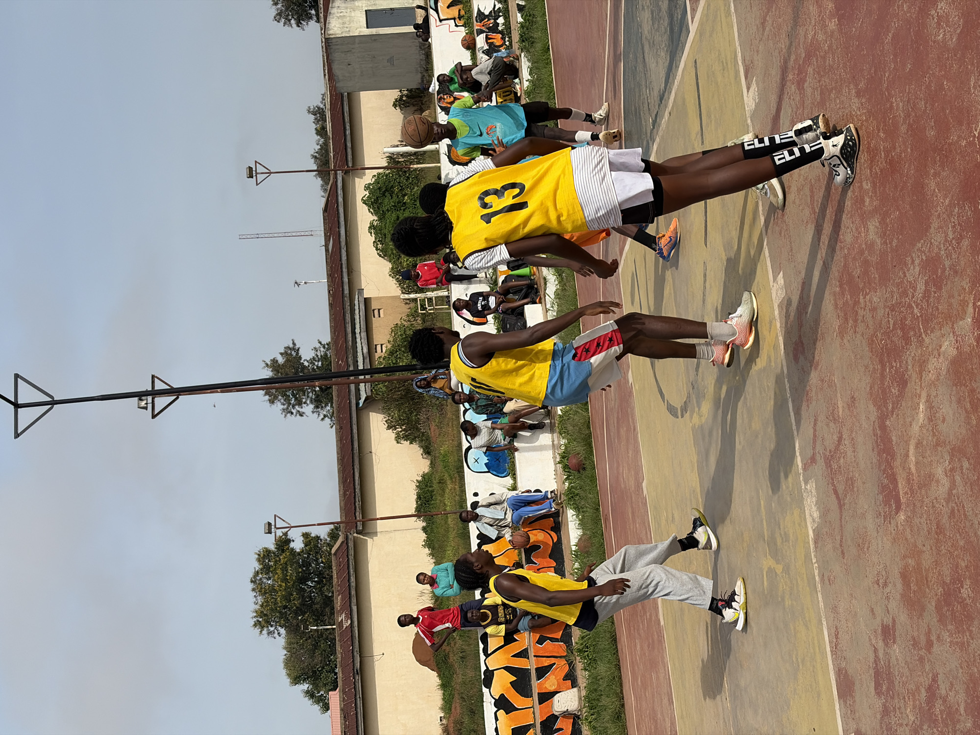 In Jos, Nigeria, a group of young people are engaged in a basketball game on an outdoor court. The court's surface is a mix of worn red and yellow concrete, with painted white lines indicating boundaries. A weathered concrete wall with vibrant graffiti art runs along the perimeter of the playing area. Spectators are seated on benches and the wall, observing the game. The background features buildings with corrugated iron roofs and lush green trees, suggesting a warm, possibly humid, climate. The sky is partly cloudy. The players are dressed in athletic wear, with some wearing jerseys numbered 13 and other identifying markings. The game appears to be in progress, with players dribbling, passing, and positioning themselves on the court.