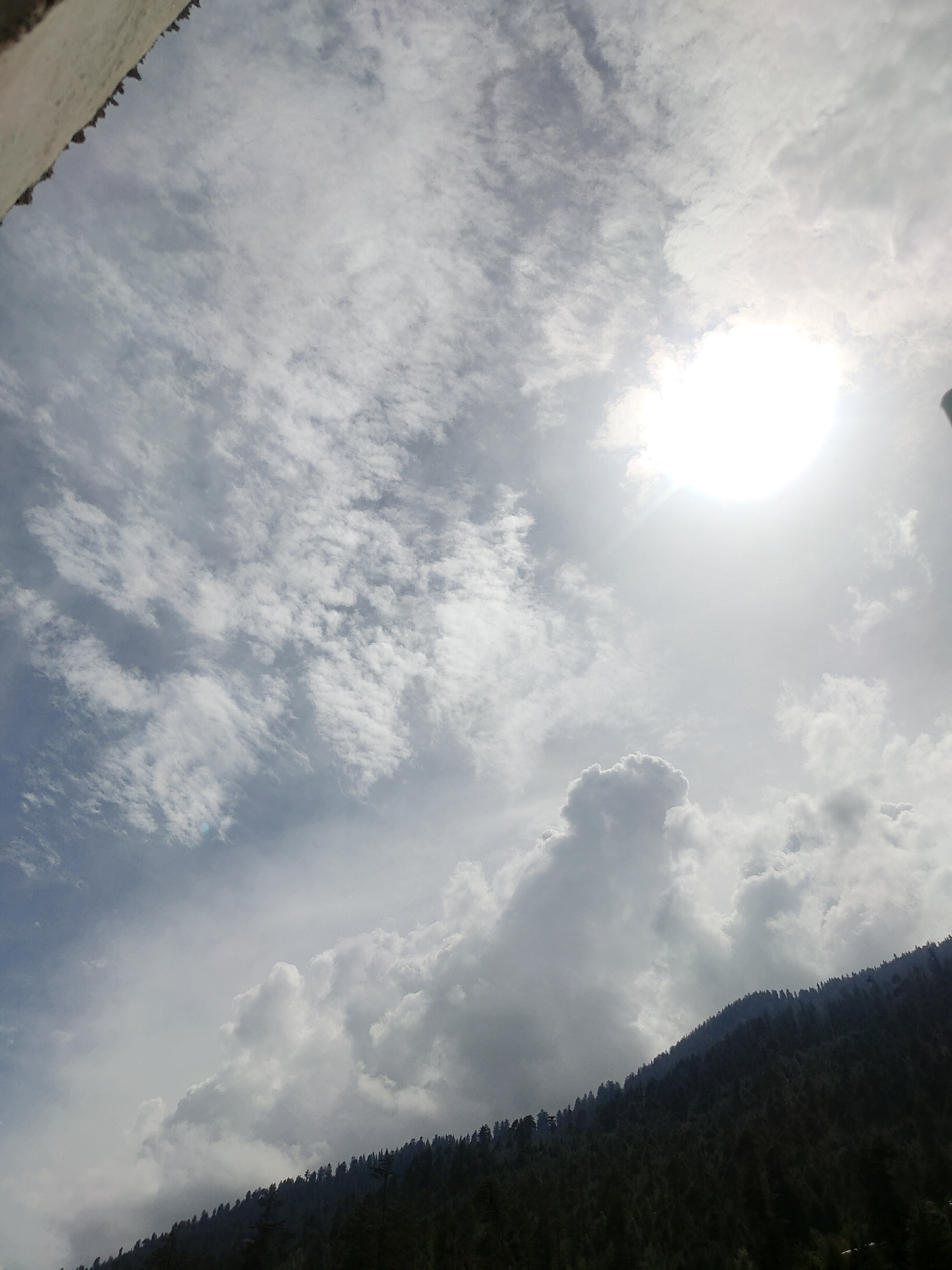This is a vertical, low-angle shot of a sky with scattered clouds and the sun. The sun, located in the upper right quadrant, is a bright, hazy orb. A sliver of a textured, light-colored building or structure is visible along the top left edge of the frame. The lower portion of the image features a dark, tree-covered mountainside. The trees are dense and form a dark silhouette against the lighter sky. The overall impression is of a natural landscape under a bright, cloudy sky, possibly taken from an elevated viewpoint. The scene does not contain any people or specific identifiable landmarks within the city of Manali, India, other than its characteristic mountainous terrain.