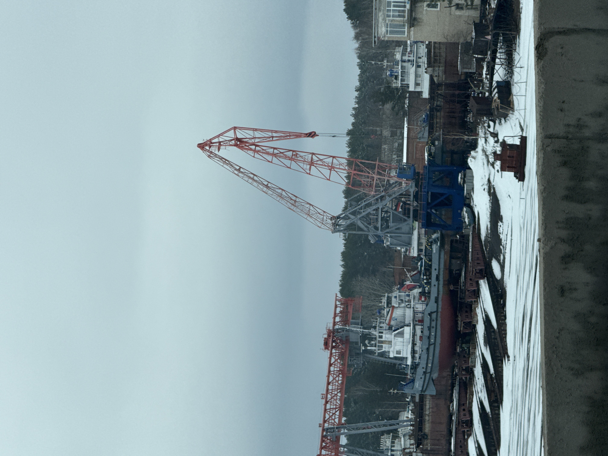 The image depicts an industrial scene in what appears to be a port or shipyard. Prominently featured is a large crane with a red lattice boom against a grey sky. The crane is situated near docked ships and various industrial equipment covered in snow. A dense tree line is visible in the background, and a building is partially visible on the right side of the picture. The photograph appears to be taken from an elevated position, possibly a building or structure overlooking the industrial area. Based on information in the prompt, the location is Chaikovsky, Russia.