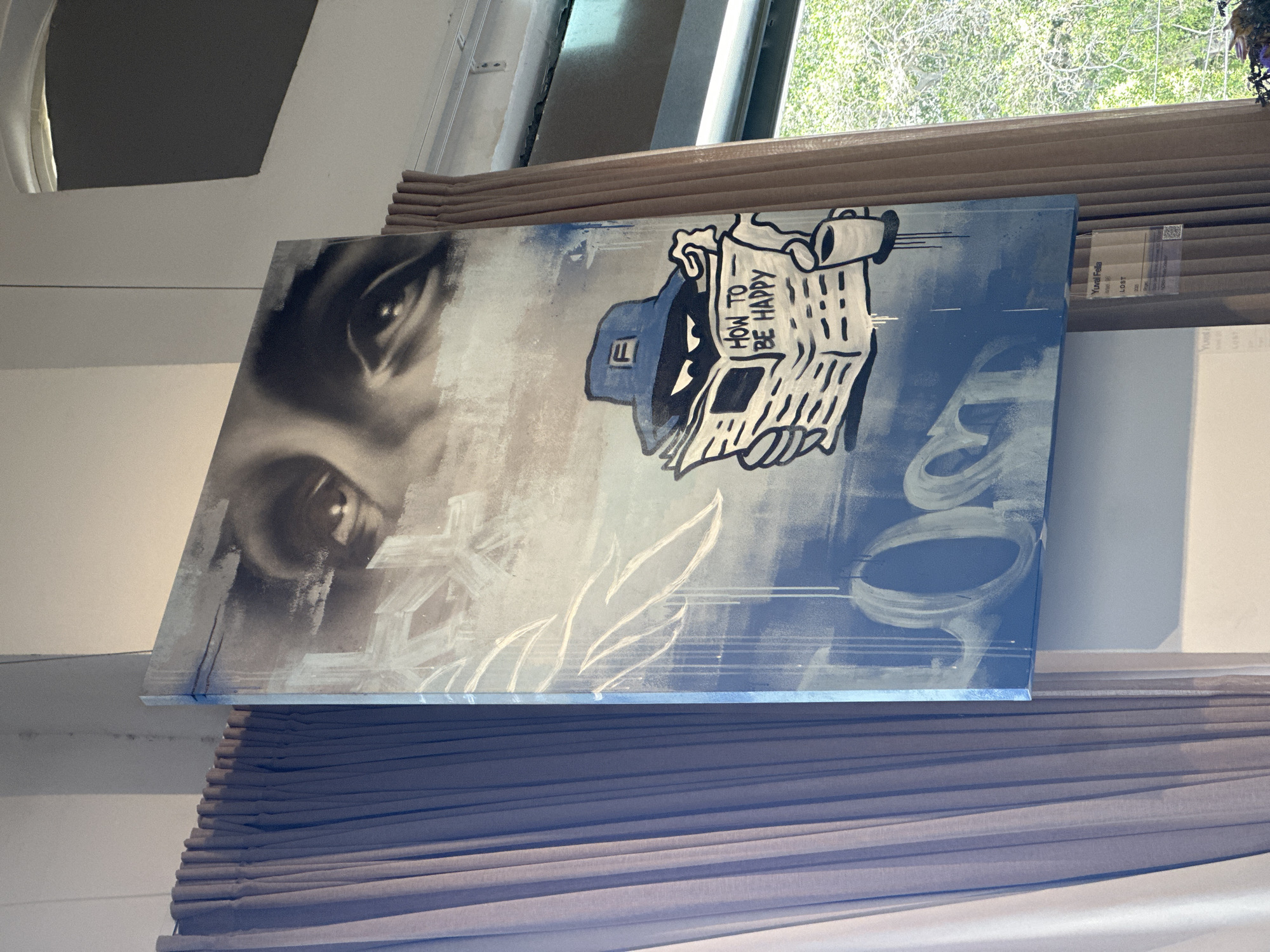 The image shows an artwork leaning against a window blind. The artwork features a close-up of a pair of eyes on the left and a cartoon-like figure holding a newspaper with the words "HOW TO BE HAPPY" printed on it on the right. The overall color palette is bluish-gray. The sticker with a title "Yuval Fella" and the word "LOST" is visible at the top right of the image. The location is likely in Tel-Aviv, Israel.