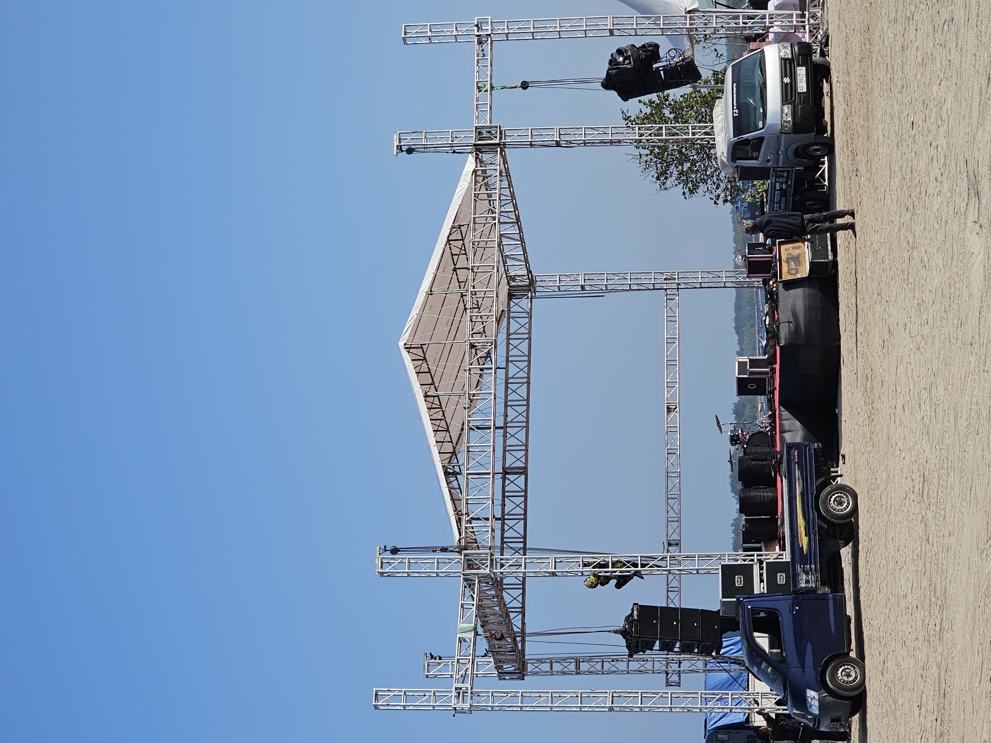An outdoor stage setup on a sandy beach with a clear blue sky above. The stage is constructed from silver metal trusses and is covered with a white tarp. Speakers are suspended from the truss structure on both sides of the stage. A blue pickup truck is parked to the left of the stage, loaded with speaker equipment. To the right of the stage, a silver pickup truck is parked. Several people are visible in the scene, some near the vehicles and others on or around the stage. In the background, there are glimpses of trees and a body of water, suggesting a coastal location. The scene appears to be a preparation for an outdoor event. The presence of stage equipment and vehicles indicates an ongoing setup process.