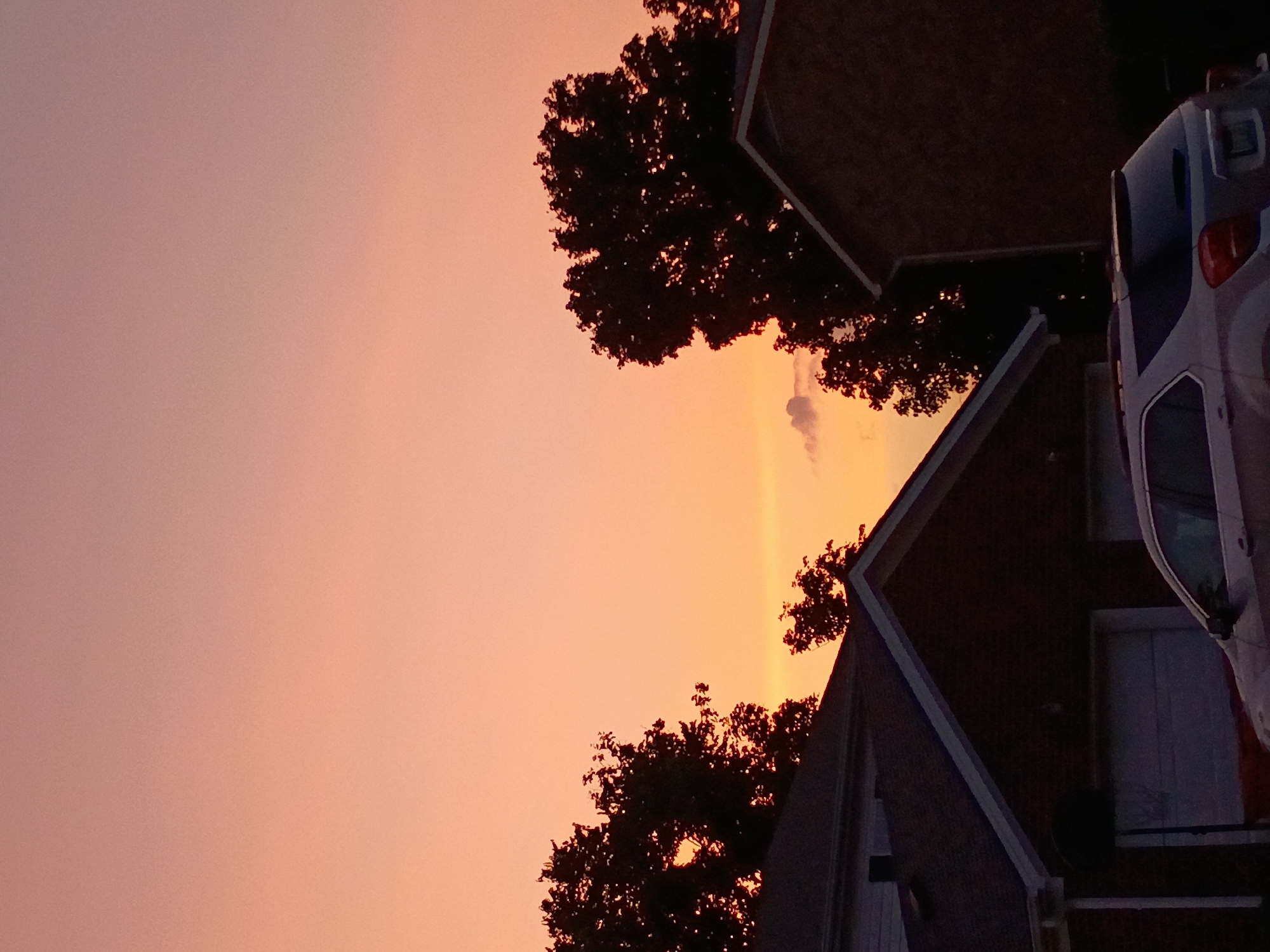 The image depicts a twilight scene with a gradient sky of pale orange and pink hues, indicative of sunset or sunrise. Silhouetted against the sky are the dark outlines of several deciduous trees and the rooftops of residential buildings. A light-colored SUV is parked in front of a garage, partially visible in the lower right quadrant of the image. The architecture suggests a suburban setting, likely in Nicholasville, United States of America, characterized by houses and garages. The overall atmosphere is peaceful and serene.