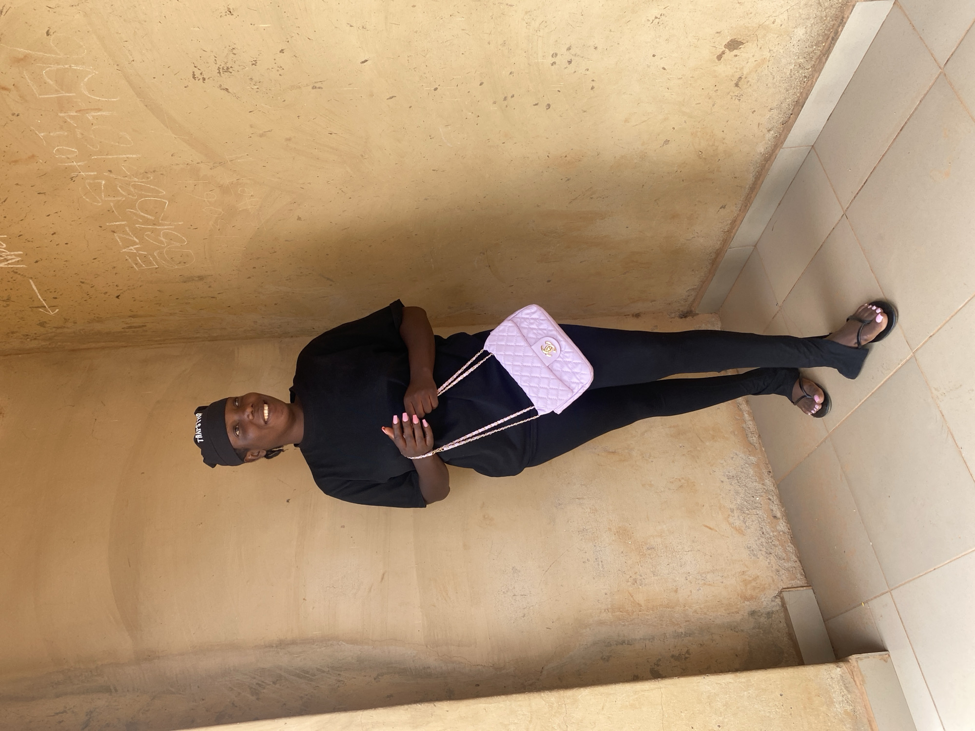 A young person stands smiling broadly at the camera in a full-body portrait, situated in what appears to be a covered corridor or sheltered walkway in Kuru Dungus, Nigeria. The subject is dressed entirely in black, wearing a t-shirt, fitted trousers, and black flip-flops that reveal pink-painted toenails. A black beanie with white text, which appears to read "TRAPSHO", adorns their head. They hold a light pink quilted handbag with a gold-colored "CC" (Chanel) logo and a chain strap, prominently displayed with pink-painted fingernails.
The background consists of a light brown, textured, and weathered wall on the left, covered with white graffiti. Visible inscriptions on the wall include "EAZI CHEF", "12", "EAZI S.B.D", "1990", and a partial "199", along with an arrow and other less discernible writings. The floor is bifurcated, with a rough, light brown concrete-like surface on the left and light beige square tiles with grey grout lines on the right. The setting appears to be a practical building environment, illuminated by natural daylight. The subject's cheerful smile conveys a positive emotion.