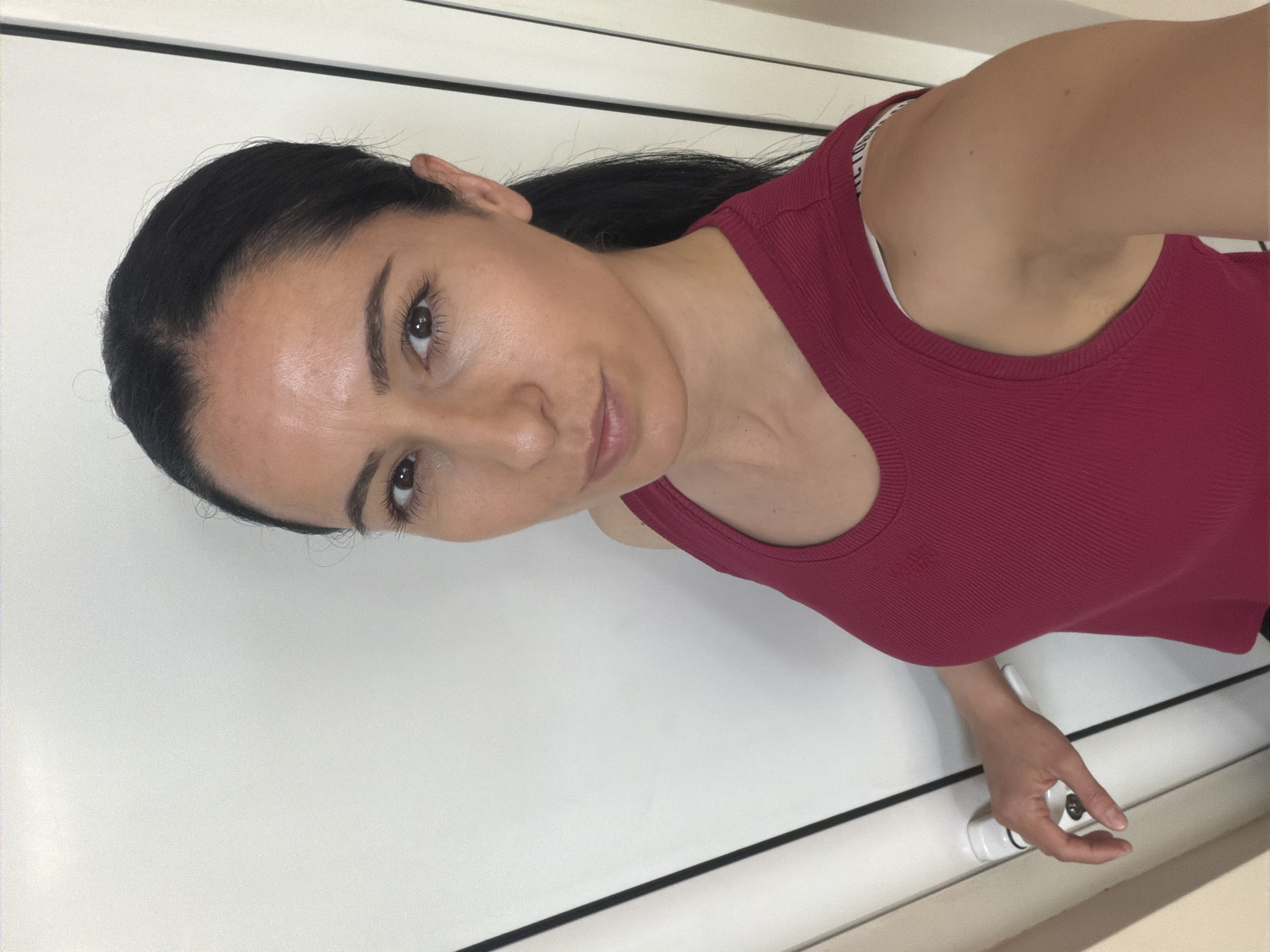 An indoor, waist-up portrait shows a woman taking a selfie, rotated 90 degrees counter-clockwise. The woman has dark hair pulled back, revealing her forehead and ears, and dark eyes looking directly at the camera with a calm, neutral expression. She wears a ribbed, dark red tank top. A white bra strap with the visible text "VICTORIA'S" is seen on her left shoulder. Her right hand, in the lower right of the frame, is interacting with a white and black latch or handle mechanism. The background consists of a plain, light-colored surface, possibly a door or wall, with a thin black horizontal line running across it. The lighting is consistent with an indoor setting. No specific location cues are visible within the image itself to confirm the provided context of Pleven, Bulgaria.
