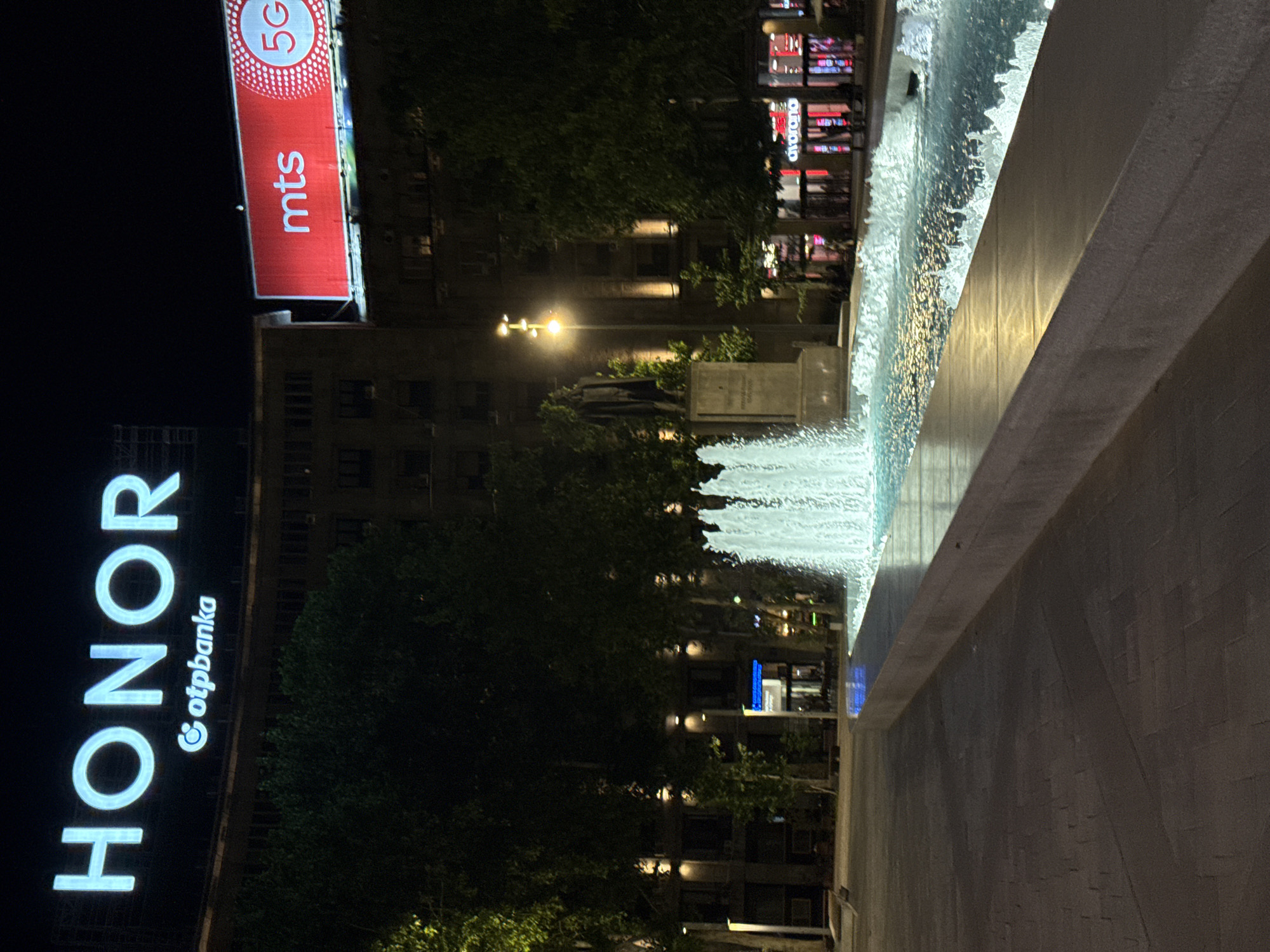 Here is a description of the image:
The image, taken at night in Belgrade, Serbia, depicts a cityscape with prominent advertisements. On the left, a brightly lit "HONOR" sign with "otpbanka" in smaller text beneath it is visible. Above and to the right of the HONOR sign, a red "mts 5G" advertisement dominates the upper left of the frame. In the background, a building with illuminated windows is seen, along with a light post and a statue. In the center of the image, there are green trees and foliage obscuring parts of the building. To the right, a fountain with flowing water is visible, with a stone structure in the foreground. The overall scene is a mix of urban elements, including commercial advertisements, buildings, and public infrastructure such as fountains.