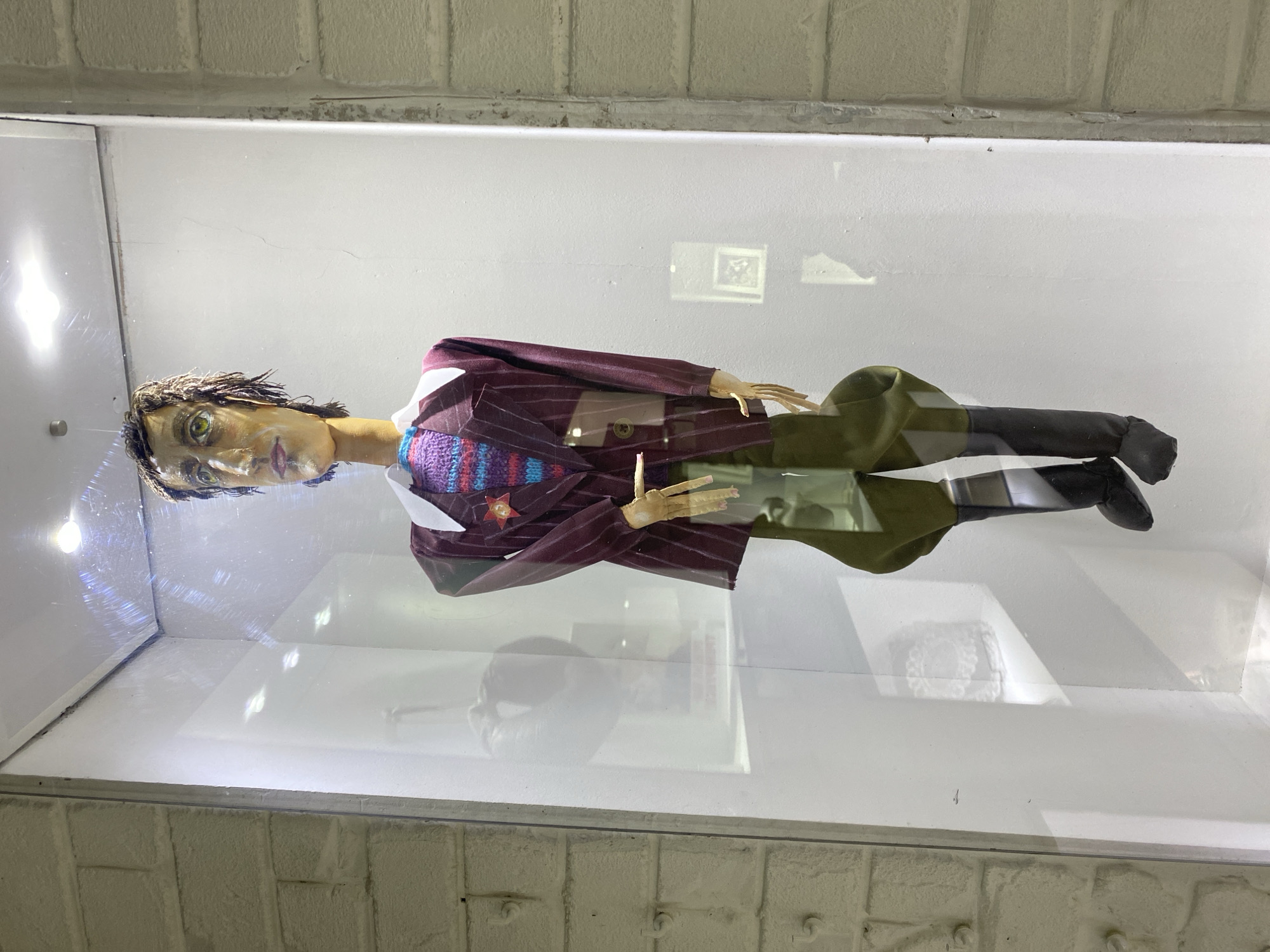 The image shows a full-body shot of a puppet displayed in a glass case. The puppet is standing upright and is dressed in a maroon pinstripe suit jacket, a multi-colored striped sweater, olive green trousers, and black boots. It has a long neck and a slender face with green eyes. The puppet is positioned in the center of the frame within a brightly lit display case. The background appears to be a white wall. The surrounding environment shows a brick wall on the left and right edges of the image. The puppet is located at the Museum of Yekaterinburg, Russia.