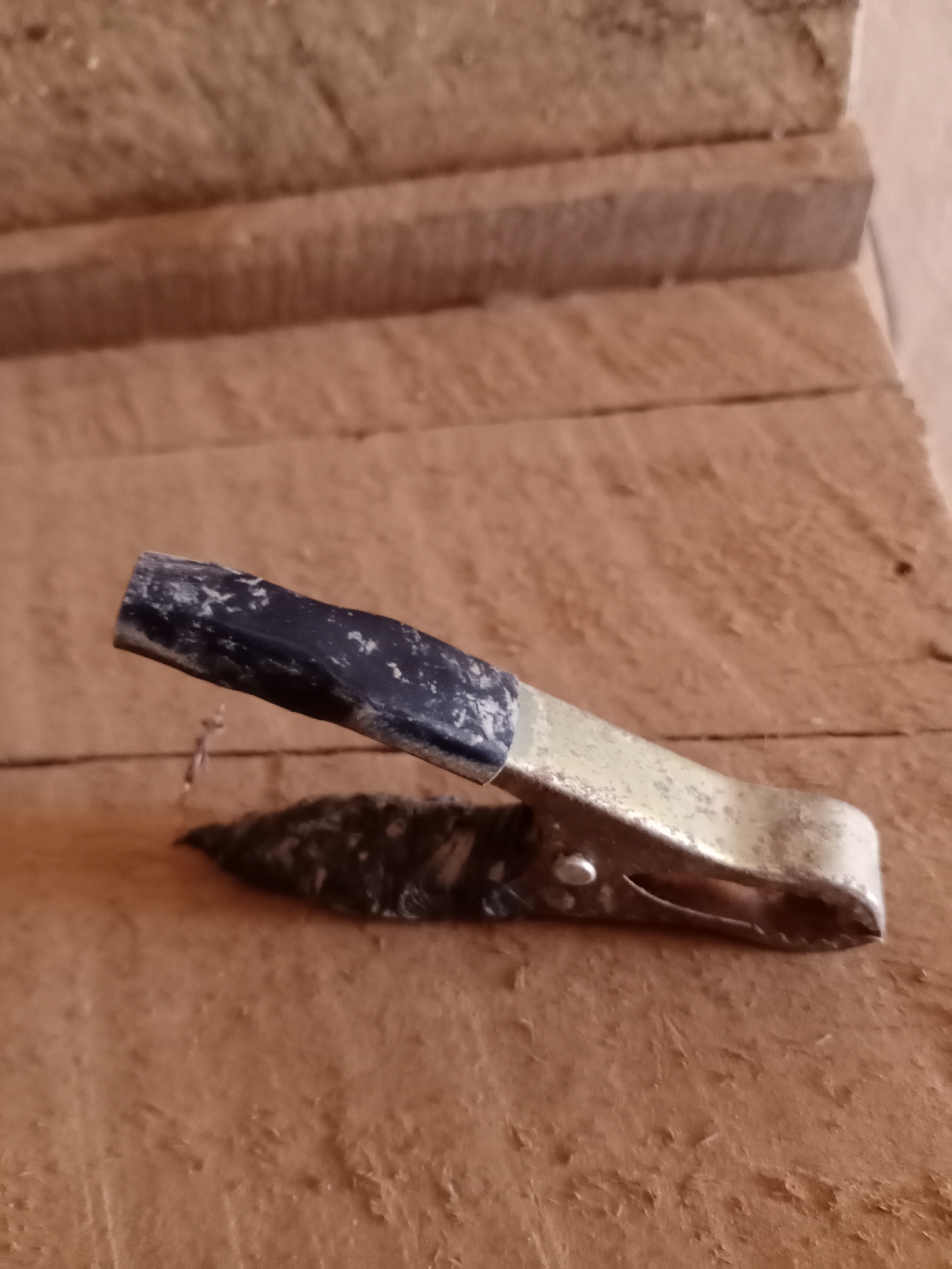 A close-up, eye-level, static shot focuses on a single alligator clamp resting on a rough, textured wooden surface. The clamp, made of metal with a black rubber grip, is angled diagonally across the frame. The metal parts of the clamp appear worn and slightly discolored, with specks of dirt or grime visible. The black grip has white scuff marks or residue. Below the clamp, a small nail is embedded in the wood. The wooden surface is composed of planks with visible grain and imperfections, suggesting an outdoor or workshop environment. In the background, more wooden planks are stacked horizontally, blurred and out of focus. The lighting is natural, casting subtle shadows and highlighting the texture of the wood and clamp. The scene is devoid of human presence or activity.