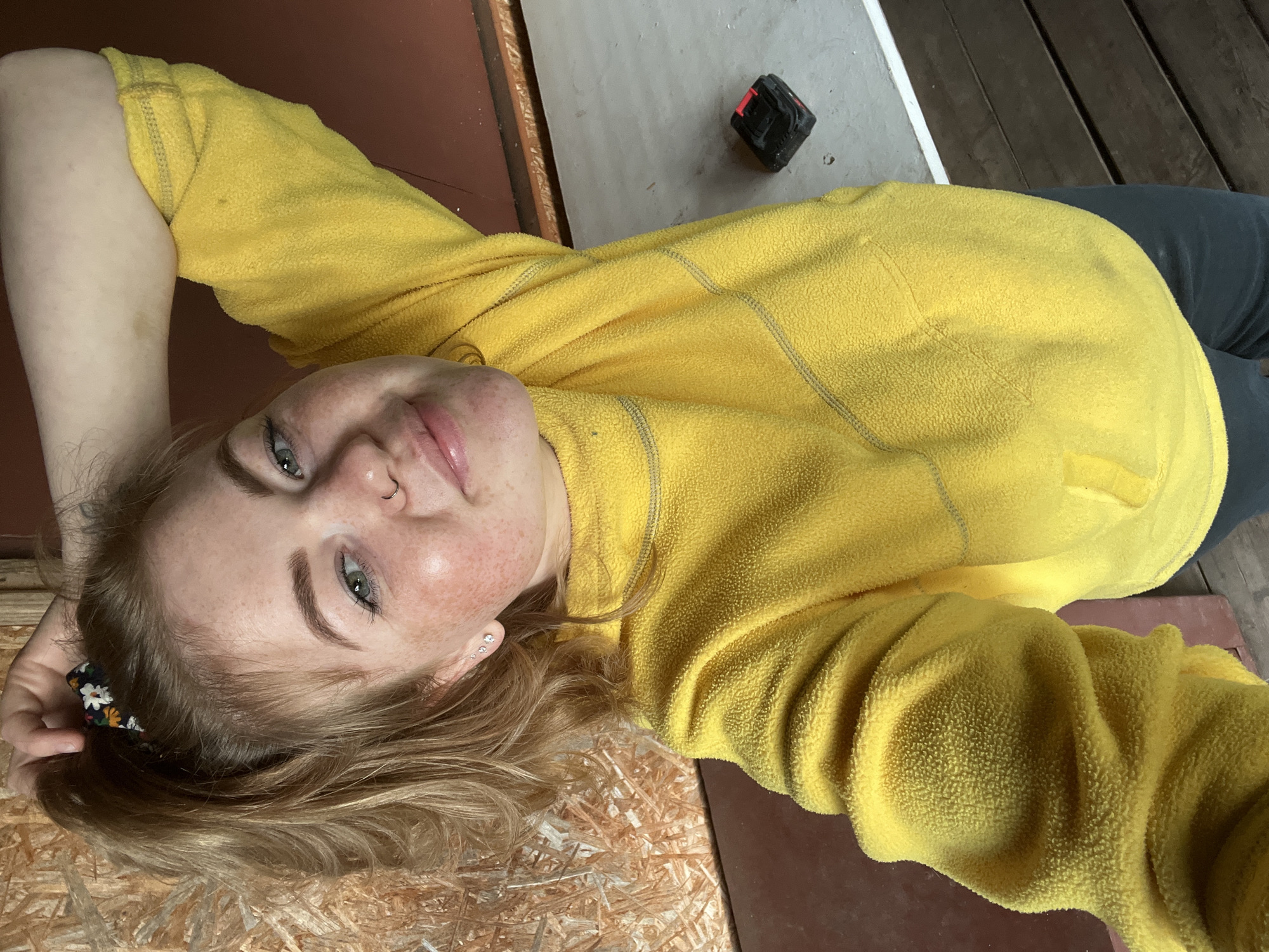 A young woman with light brown hair and prominent freckles on her face is featured in a portrait-style image, appearing to be lying down and looking upwards towards the camera with a gentle, relaxed smile. She has blue-green eyes, multiple stud earrings in her left ear, and a nose piercing. She is wearing a bright yellow fleece sweater, and a floral-patterned scrunchie is visible on her left wrist.
The setting appears to be an indoor or semi-enclosed space in Chaikovsky, Russia, characterized by varied floor surfaces. Directly beneath her head is a light-colored wooden board, possibly OSB. To her right, a reddish-brown surface is visible. Further into the background, the floor transitions to a light grey concrete or painted area, which then meets dark wooden planks. A compact black and red object, resembling a small tool or tape measure, rests on the grey surface. The overall atmosphere is casual, and the lighting suggests daytime. There is no visible text in the image.