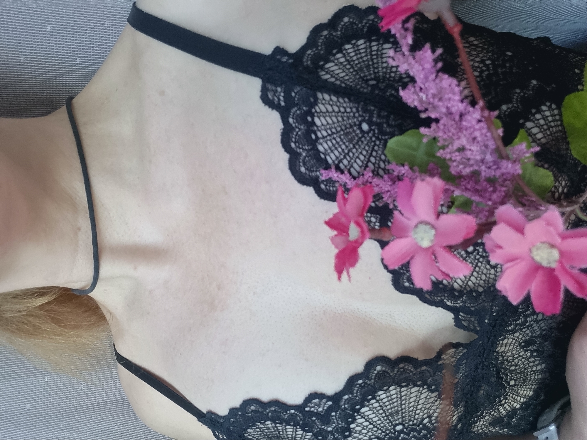 The image presents a close-up view of the upper torso of a person, likely female, captured from an overhead perspective. The subject is wearing a black lace lingerie garment, which features intricate floral patterns, and a thin black choker necklace around their neck. A slender black strap from the garment crosses the collarbone.
A small, artificial floral arrangement is placed on the lace garment on the right side of the frame, consisting of several bright pink daisy-like flowers with distinct white centers, alongside some fuzzy purple spike-like flowers and green foliage. These flowers add a pop of color against the dark lace and fair skin.
The person's fair skin shows minor variations in tone and texture. A hint of blonde hair is visible near the left shoulder, suggesting the subject has light-colored hair. The individual appears to be reclining on a textured light grey or off-white fabric surface, possibly a bedspread or sofa, which forms the background of the shot. The lighting appears soft and natural, indicating an indoor setting, likely with ambient daylight.
The image emphasizes the aesthetic contrast between the soft skin, the delicate black lace, and the vibrant artificial flowers. The composition suggests a posed portrait or fashion shot. There is no discernible activity beyond posing for the photograph. No visible text is present in the image. While the context states the location is Chaikovsky, Russia, the image itself provides no visual cues to confirm this geographical detail.