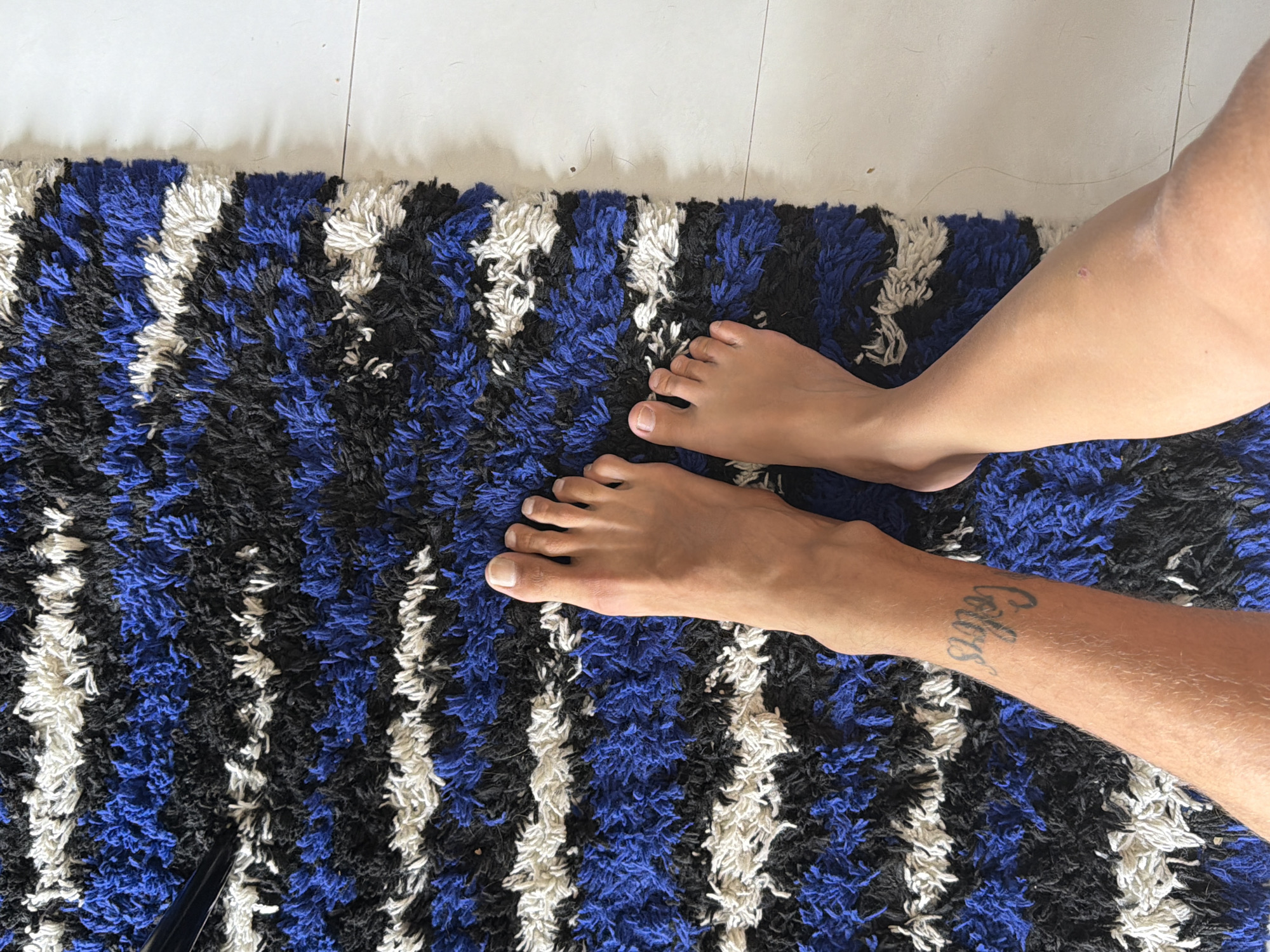 The image shows a partial view of a person's legs and feet resting on a textured rug. The rug has a repeating striped pattern consisting of blue, black, and white hues. The person's left leg is visible, displaying a tattoo on the lower portion of the leg near the ankle. The person's feet are bare. The rug is positioned on a white tiled floor. The scene's location within the city of Chang Phueak, Thailand, cannot be determined from the visual information provided.