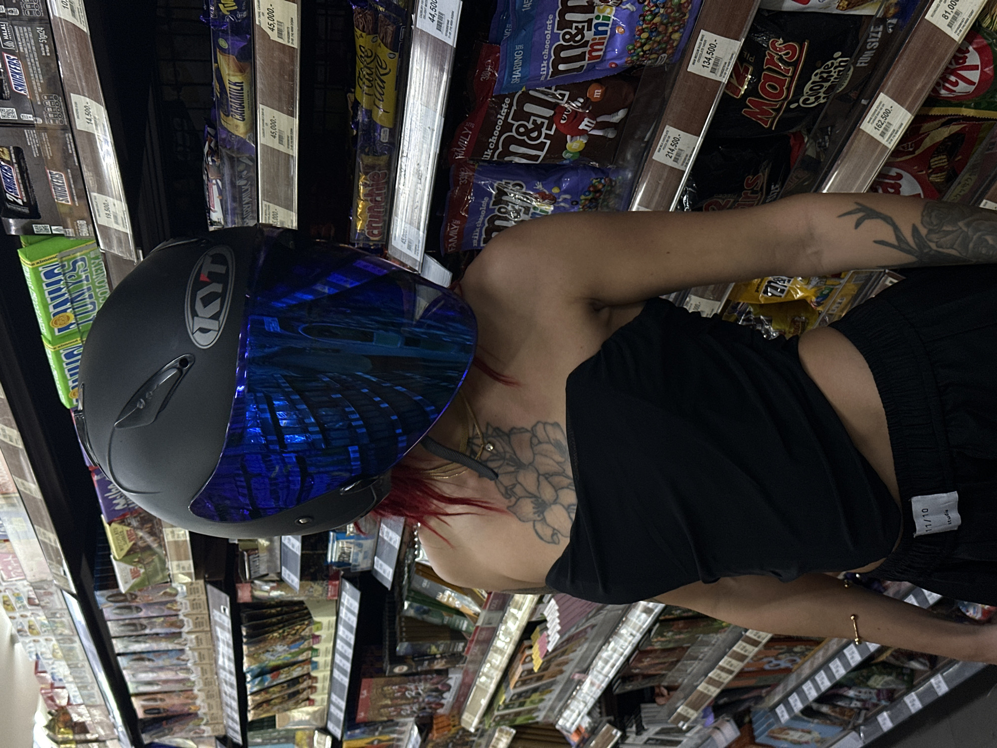 A person wearing a black strapless top and black shorts stands in a grocery store aisle filled with candy. The person is wearing a black motorcycle helmet with a blue reflective visor. Their back and arm are heavily tattooed. Candy bars such as Snickers, Kit Kat, and Mars are visible on the shelves. The candy bars display prices in Indonesian Rupiah. This image appears to be taken in a grocery store in North Kuta, Indonesia.