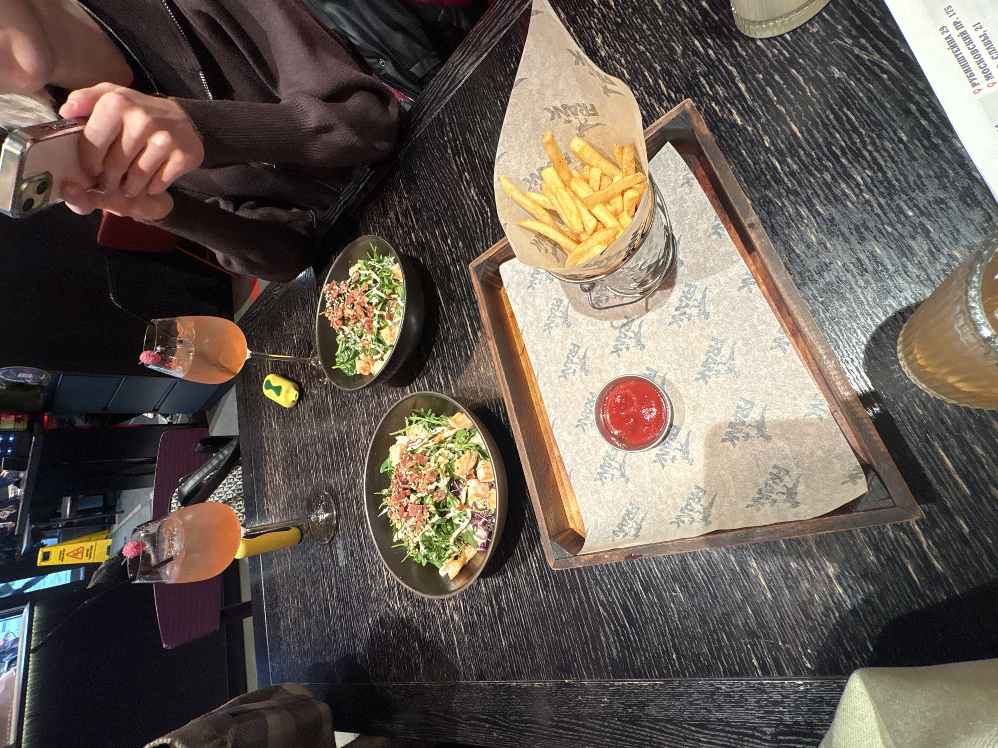 The image shows a restaurant table from above, featuring a brown wooden surface with a dark, textured finish. The table is set with two bowls of salad, a metal container of french fries in a paper cone, a small dish of ketchup, and two cocktails in stemmed glasses. A hand holding a cell phone is visible in the upper left corner, suggesting someone is taking a picture. The food is presented on a wooden tray lined with paper printed with the word "FRANK." A drink menu is visible in the upper right. A sign in the lower left suggests construction nearby.
The OCR reveals two locations in Saint Petersburg, Russia: Rubinstein Street 29 and Moscow Avenue 175, as well as Slava Avenue 21.