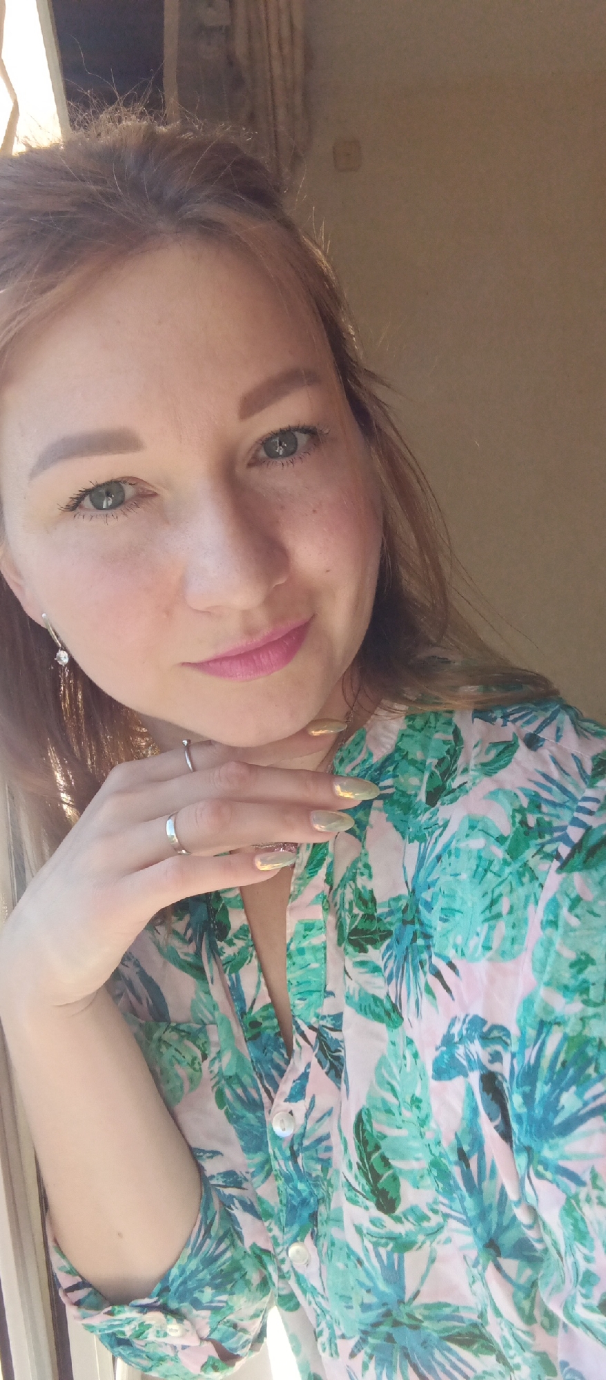 A woman is posing for a close-up selfie in what appears to be an indoor setting. She is wearing a tropical-themed button-down shirt in shades of green, blue, and pink. She has fair skin, light brown hair with blonde highlights, green eyes, and pink lipstick. She is wearing rings on her left hand, which is posed with her fingers resting on her chin. She also has earrings and her nails are painted with a pearlescent finish. The background is a plain wall with a window on the left side of the frame. The location is Unknown, Unknown.