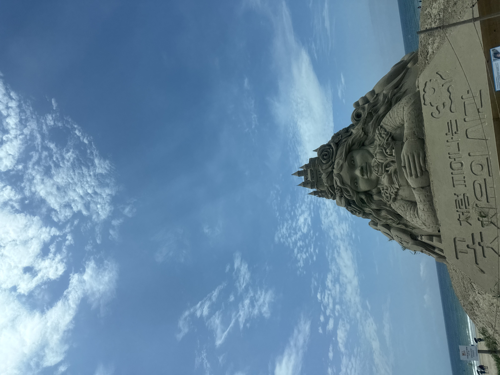 The image shows a sand sculpture of a woman with a castle on her head, located on the beach in Anmyeon-eup, South Korea. The sculpture is detailed, with the woman having long, flowing hair, and she is holding a bouquet of roses. The castle is intricate, with towers and crenellations. The sculpture is placed in front of the ocean, with the blue sky and white clouds forming the backdrop. There are some people in the distance on the beach, and a signpost is visible to the left of the sculpture. The sand sculpture is a prominent feature of the scene, and the overall impression is one of artistry and natural beauty.