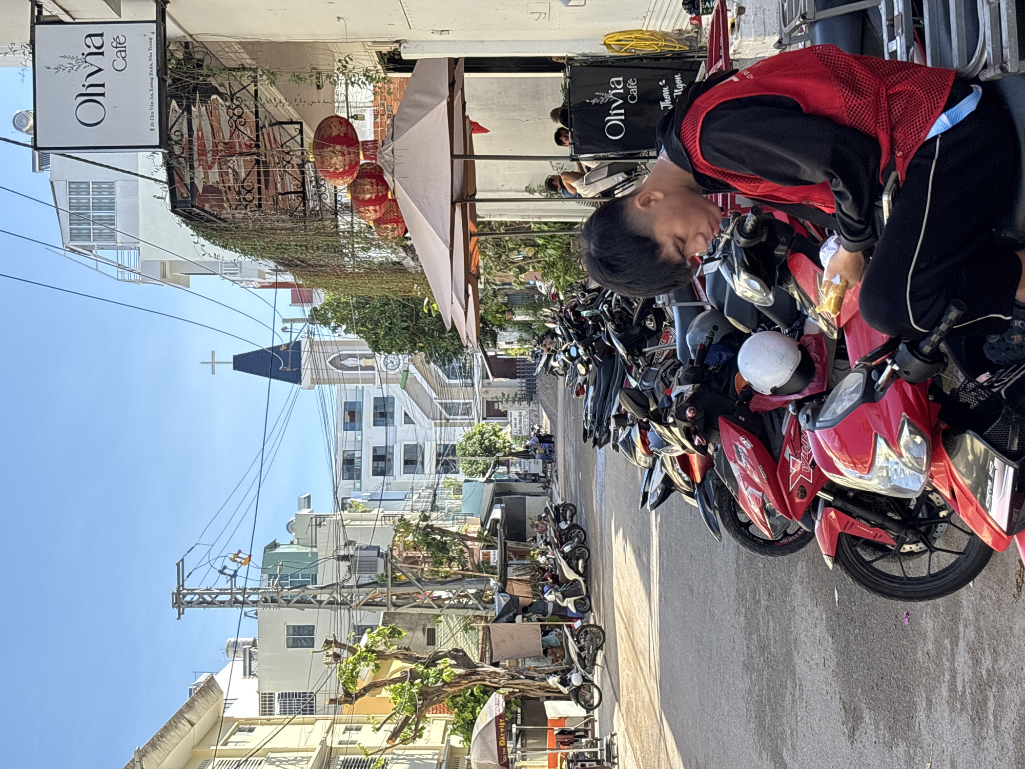 In a sunlit street in Nha Trang, Vietnam, a young man sits on a motorcycle, engrossed in his phone and a piece of food. The street is lined with parked motorcycles, and buildings rise on either side. A sign for "Olivia Cafe" hangs above an outdoor seating area, complete with umbrellas and hanging lanterns. In the background, a church steeple with a cross stands against a clear blue sky. The scene depicts a typical urban streetscape, with a mix of commercial and residential buildings, and the ubiquitous presence of motorcycles.