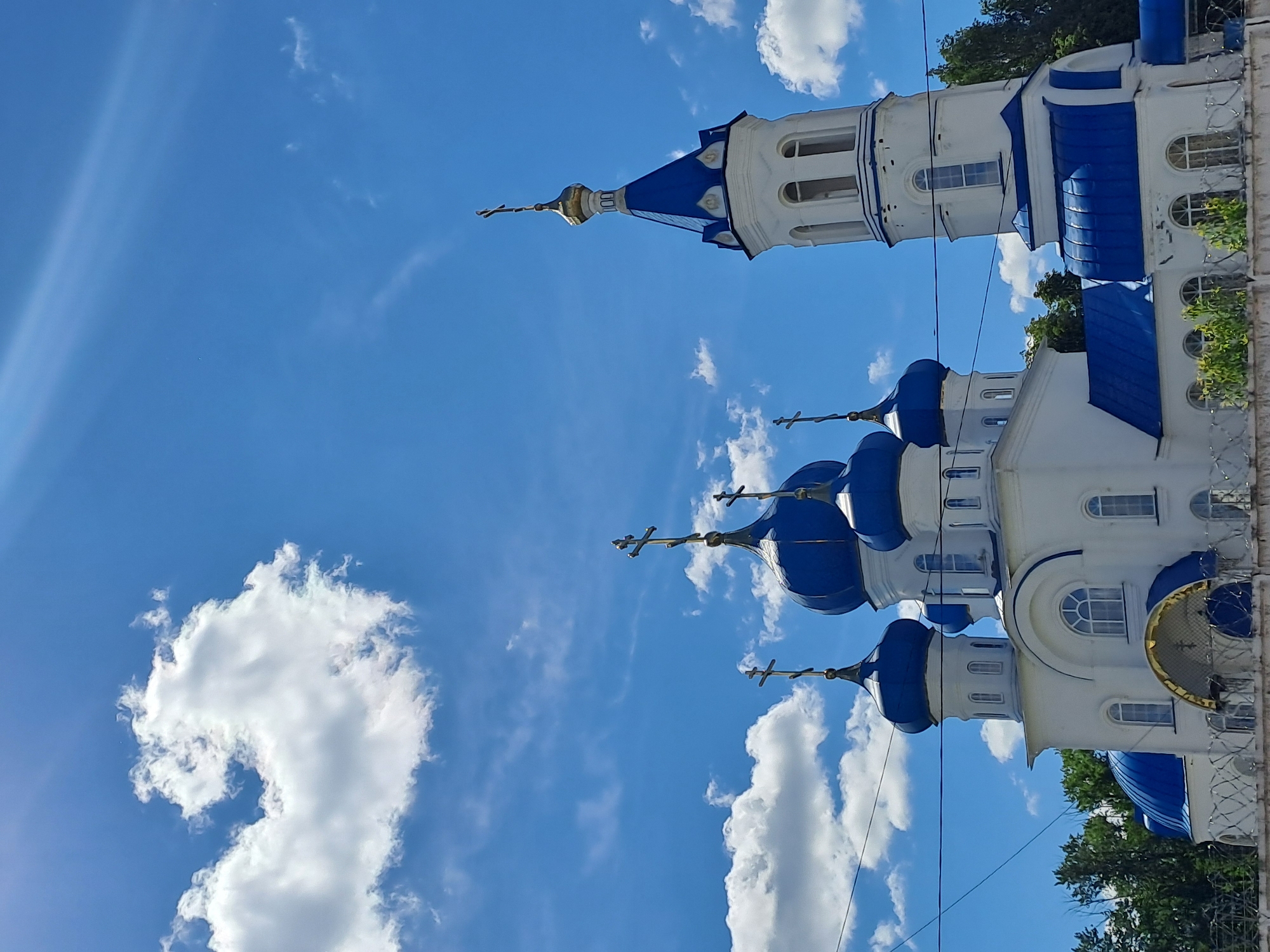 A majestic Orthodox church stands prominently against a bright blue sky. The church features pristine white walls and multiple vibrant blue onion domes, each topped with a golden cross. A tall, elegant bell tower, also crowned with a blue conical roof and a cross, is visible on the right side of the main structure. The architecture includes numerous arched windows and decorative elements.
The setting is an outdoor, sunny day, indicated by the clear, brilliant blue sky scattered with fluffy white clouds. Sunlight casts a gentle glow, and a subtle lens flare is visible in the upper left corner. Lush green trees can be seen along the right edge of the image, partially behind the church. A fence, appearing to be decorative metalwork or barbed wire, runs along the bottom right, bordering the church grounds.
No people are visible, and no specific activities are taking place; the image focuses on the church's architecture. There is no visible text. The style of the church is characteristic of those found in Russia, aligning with the given location context of Yalga, Russia.