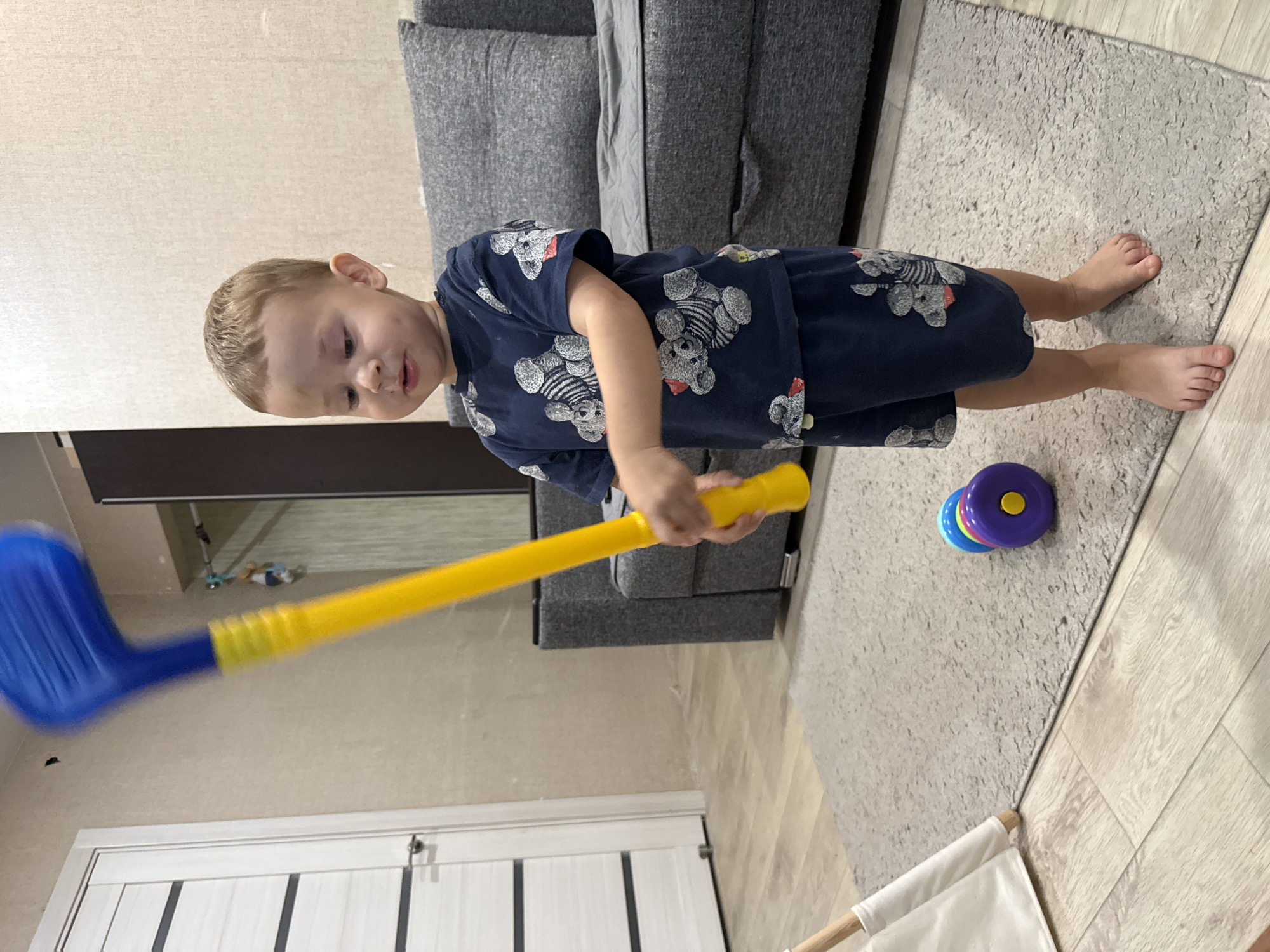 A young Caucasian boy, appearing to be around 2-3 years old, stands barefoot on a grey rug in what appears to be a residential living room. He is wearing a dark blue pajama set adorned with a repeating pattern of cartoonish bears. The boy is holding a large, bright yellow toy golf club with a blue head, raised as if about to swing it. He is looking down at the club with a focused expression. To his right, a grey sofa is visible, and to his left, part of a white tent-like play structure is present. On the rug in front of him, a colorful toy stacking ring with blue, purple, and yellow rings and a yellow knob is lying on its side. The background shows a neutral-colored wall and a door with horizontal panels. The overall lighting suggests an indoor environment, possibly during daytime. The location is specified as Rusanovo, Russia.
