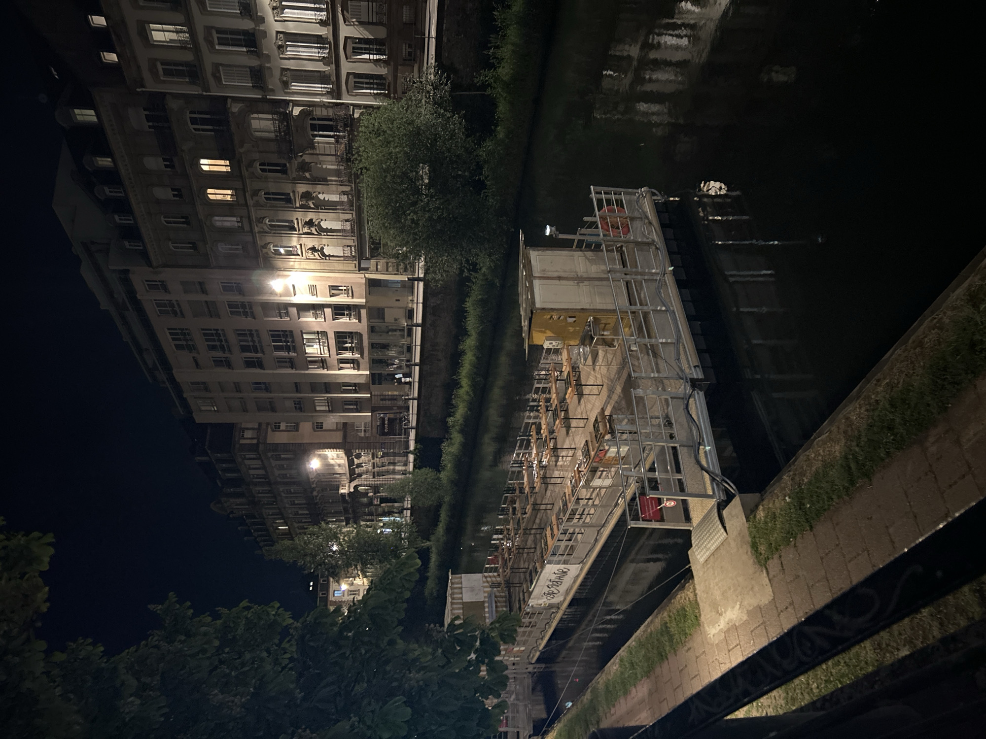 The image depicts a nighttime scene along the canals of Strasbourg, France. Multi-story buildings with numerous windows and classical architectural details are visible to the left of the frame, illuminated by streetlights and some interior lighting. A tree line and vegetation mark the canal's edge along the buildings.
In the canal, a large, multi-level floating structure is moored to the side. The structure appears to be a restaurant or bar with numerous tables and chairs. It is constructed with a scaffolding-like frame and features various levels and platforms. Signage is visible on the side of the floating structure.
The canal's water surface reflects some of the surrounding lights. The opposite side of the canal features a brick walkway and grass, with a dark foreground element possibly being a fence or railing.
The scene is dimly lit, contributing to a somewhat mysterious atmosphere. There is no indication of people within the frame.