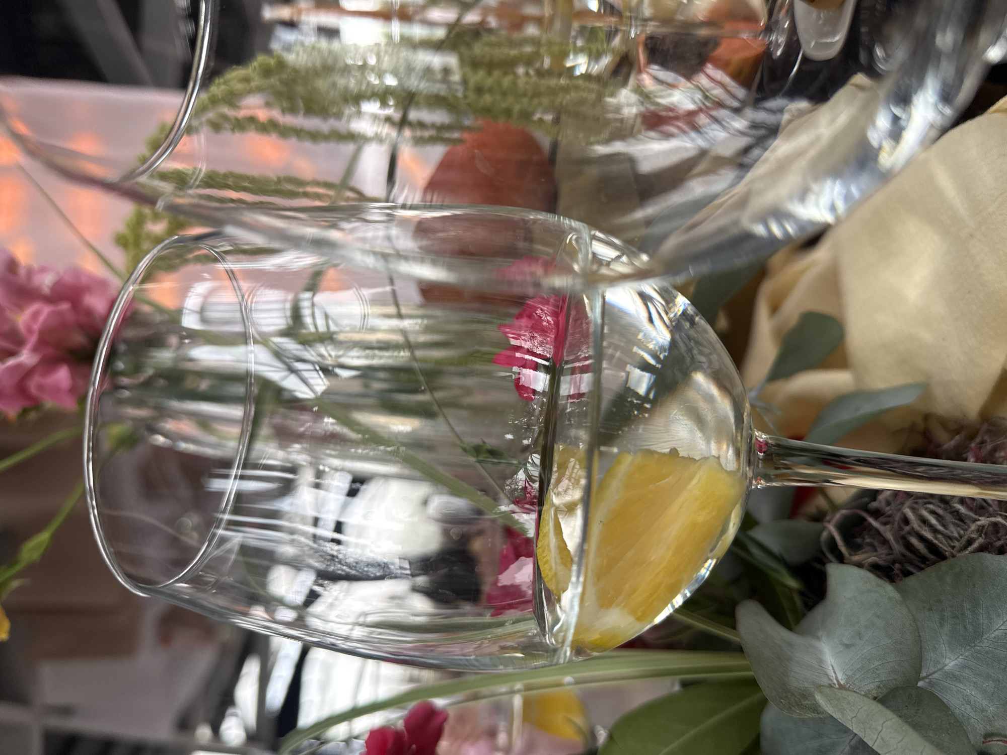 The image presents a close-up view of a clear, empty wine glass with a slice of lemon inside, positioned amidst an arrangement of flowers and greenery. Pink and red flowers are visible, along with eucalyptus leaves. The scene has elements of a decorative or event setup. The location is not discernible from the image.