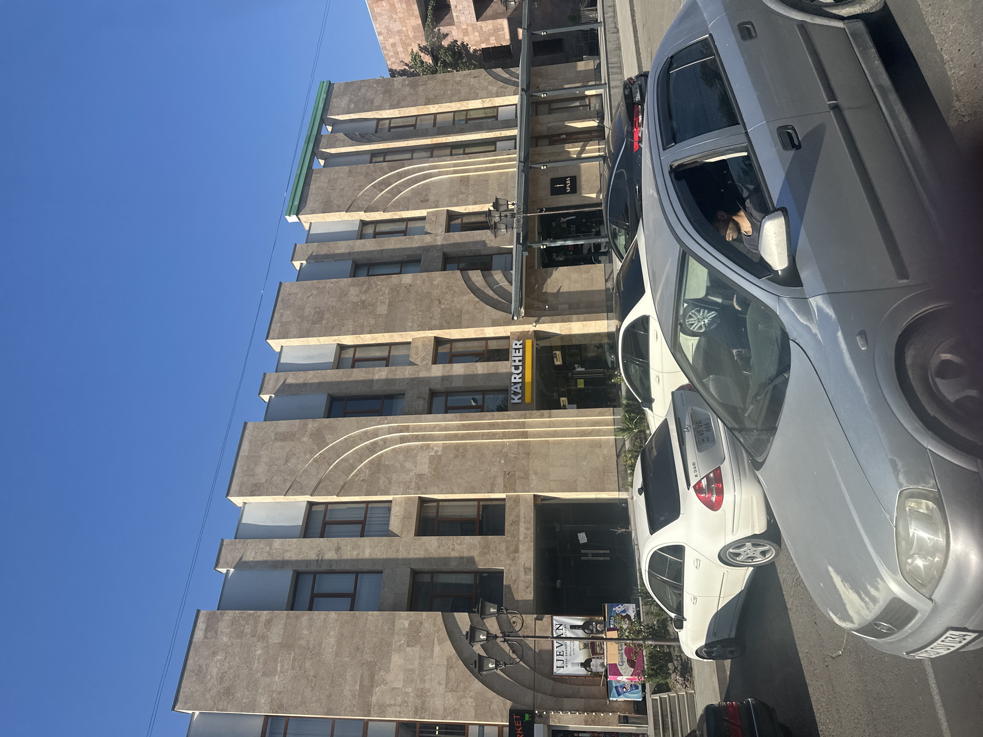 The image shows a street view in Gyumri, Armenia, featuring a modern building with a beige stone facade and distinct vertical and arched architectural elements. The building has multiple windows and signs, including "KARCHER". In the foreground, several cars are parked or driving on the street. A silver car is prominently displayed with a man inside. To its left, a white Mercedes sedan is visible, and further left, a black sedan. The sky is clear and blue. A street lamp with multiple lights stands near the building entrance. Advertisements for "IJEVAN WINE BRANDY FACTORY" are also present. The scene appears to be a commercial or office building area.