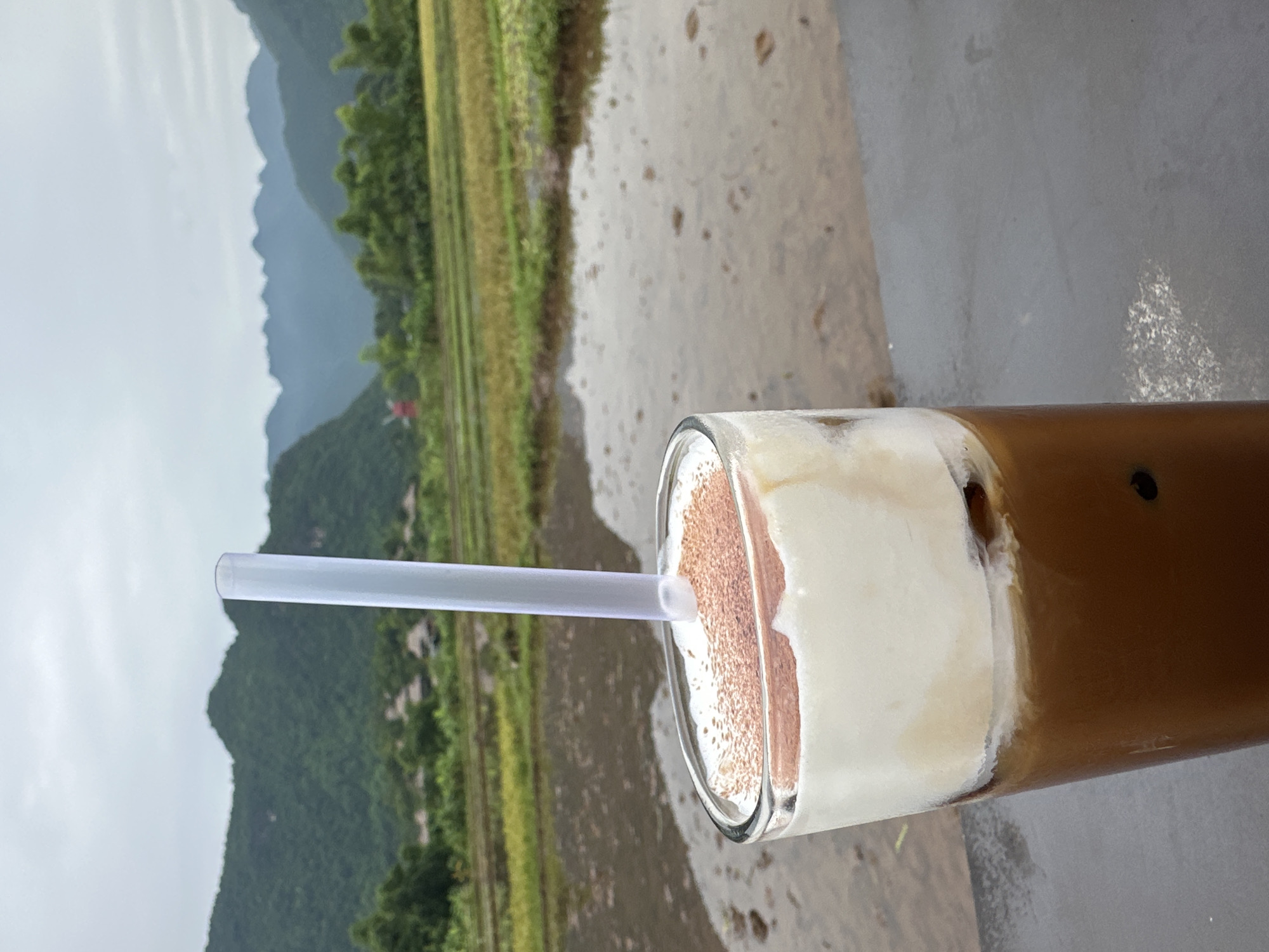 A tall glass of iced coffee with a layer of white foam topped with cinnamon powder is in the foreground, positioned on a dark surface. A white plastic straw extends from the drink. Behind the glass, a body of water reflects the sky and the surrounding landscape. The middle ground features a rice paddy with green and golden stalks. In the distance, lush green mountains rise against a cloudy sky. A small red windmill is visible among the trees on the left side of the frame. The scene is set in the rural outskirts of Mai Chau, Vietnam, likely a scenic area known for its rice terraces and mountainous terrain.