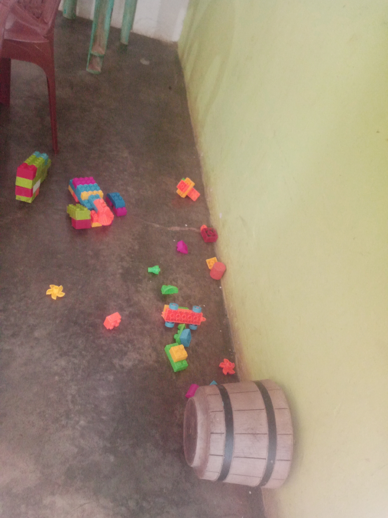 In La Tagua, Venezuela, a dimly lit scene unfolds on a worn concrete floor. Scattered across the floor are various colorful children's building blocks, some assembled into small structures. Among these are pink, blue, green, yellow, and orange plastic blocks, with one larger, red and green structure prominently positioned to the left. Further to the right, a miniature wooden barrel with dark bands sits on the floor, its end facing the viewer. The background consists of a pale yellow wall, its texture slightly uneven, and the legs of two chairs are visible on the left side of the frame, one a dark red plastic chair and the other appearing to be green metal. The lighting is muted, casting soft shadows.