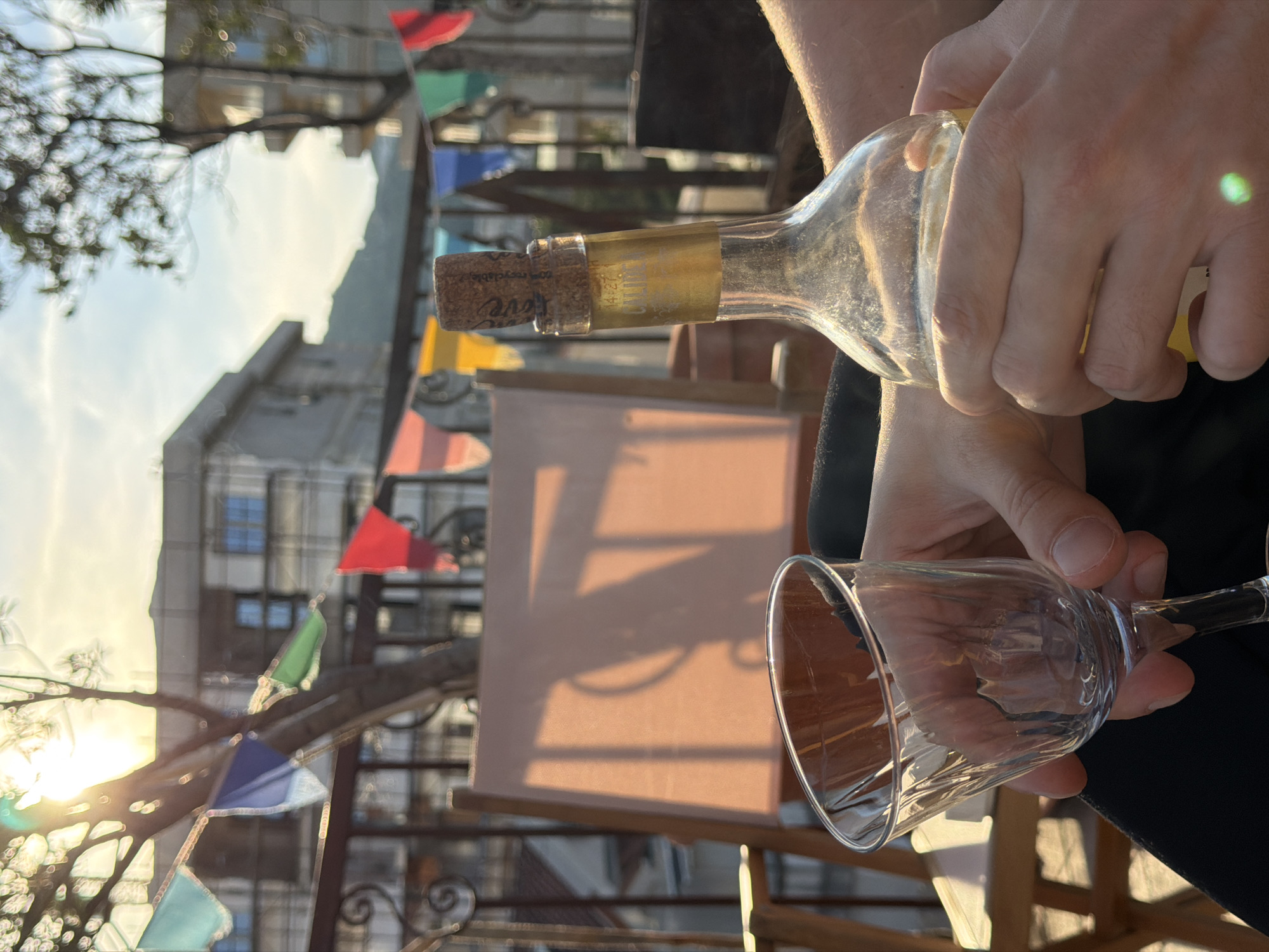 The image shows a person pouring white wine from a bottle into a glass. The cork is in the bottle. The scene is likely outdoors, possibly on a terrace or balcony, given the presence of sunlight, a tree branch, and colorful pennant flags in the background. A building is partially visible in the distance. The location appears to be in Budva, Montenegro, based on the architectural style and the presence of Adriatic Sea vegetation.
