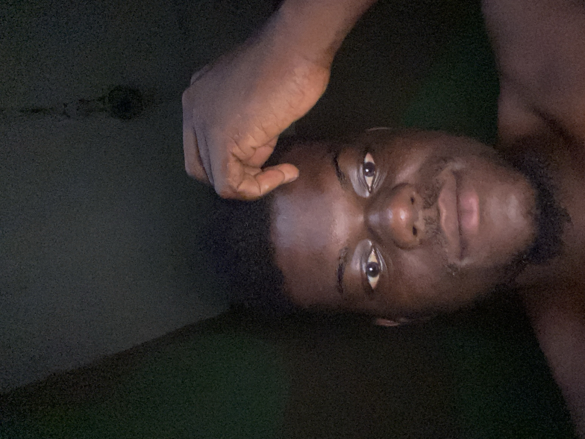 A dark-skinned man is shown from a close-up, angled perspective, with his face positioned towards the right. His hand is raised to his forehead. The background is a dimly lit, textured surface. The city and location are unknown.
