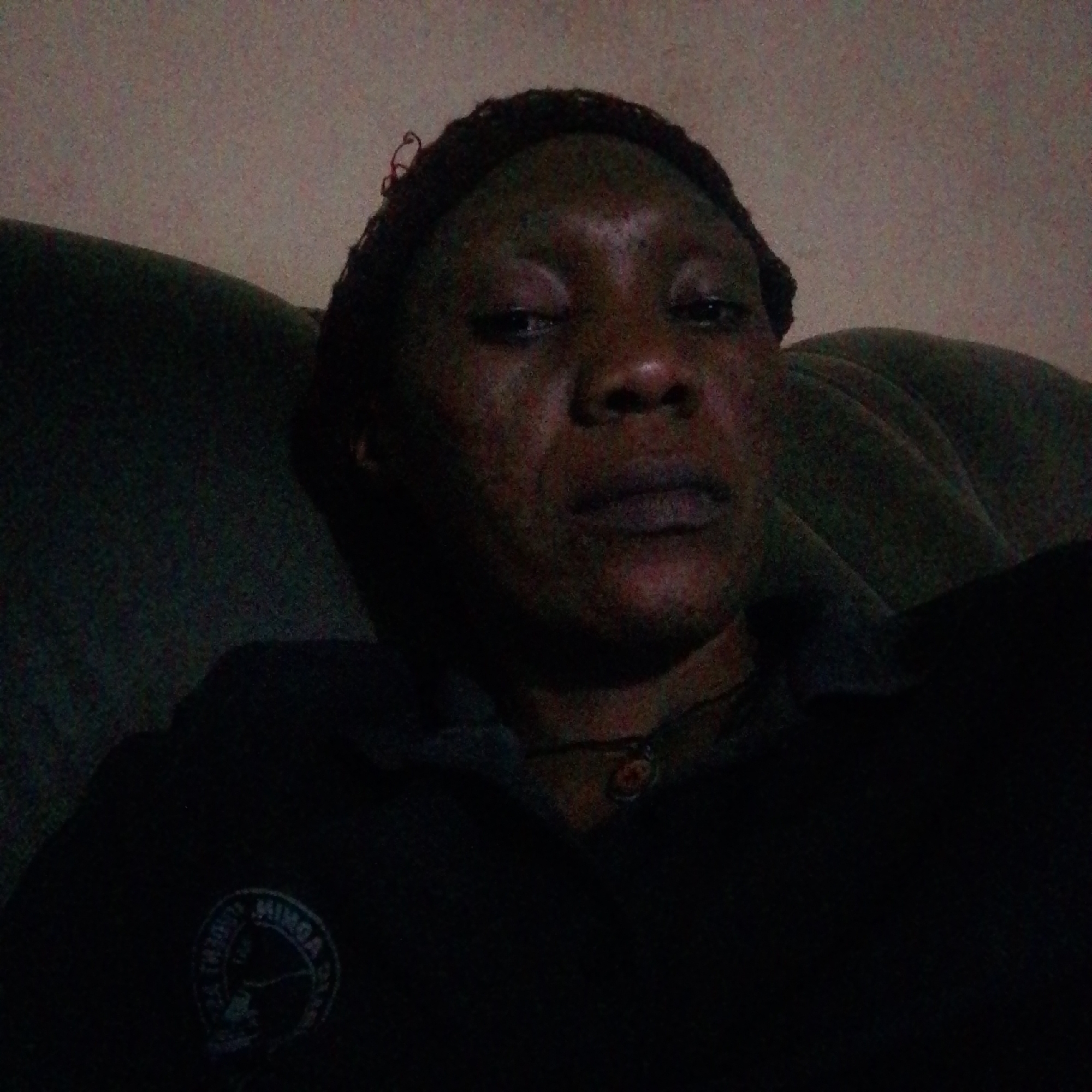 A person with dark skin and short, dark hair sits on a green, upholstered couch. The person wears a dark shirt with a circular logo on the left breast. The photo was taken indoors in dim lighting. Location: Unknown, Unknown.