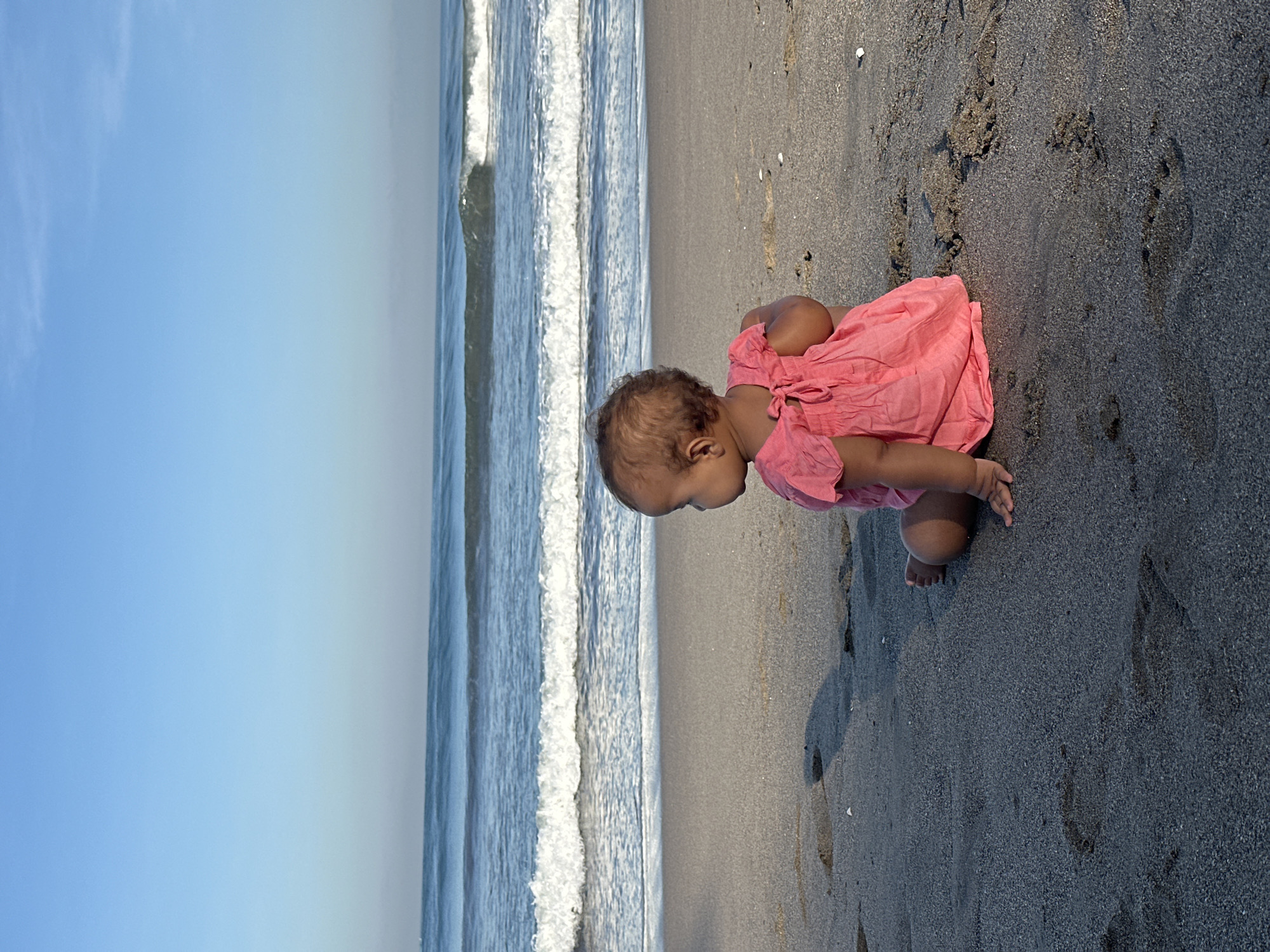A baby is sitting on a beach in North Kuta, Indonesia, facing away from the camera and looking out at the ocean. The baby is wearing a pink dress with a bow in the back. The sand is dark and wet, with footprints visible. The ocean has gentle waves and a clear blue sky above.