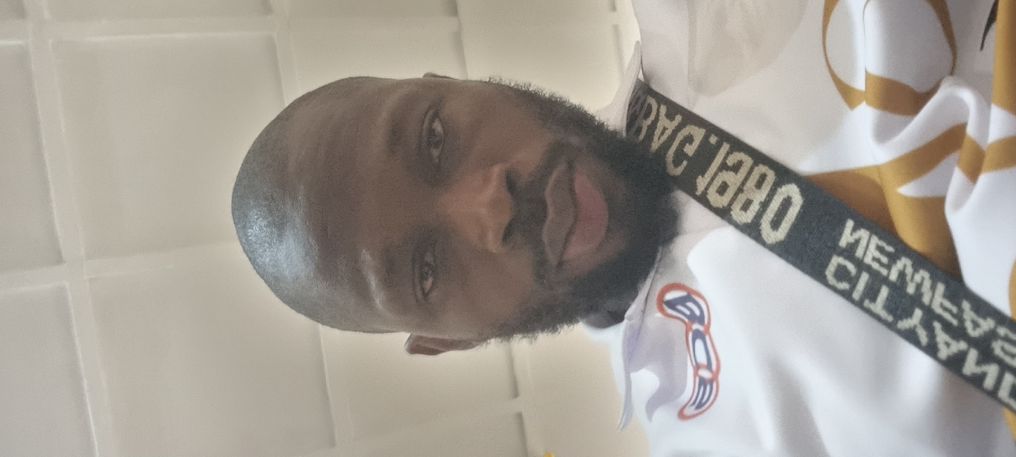 A portrait of a dark-skinned man with a shaved head and a beard. He is wearing a white shirt with a brown pattern and a black strap with the words "ABEL 1980" and "MEMBER CITYLAND" printed on it. The background consists of white tiles. The location is Unknown, Unknown.