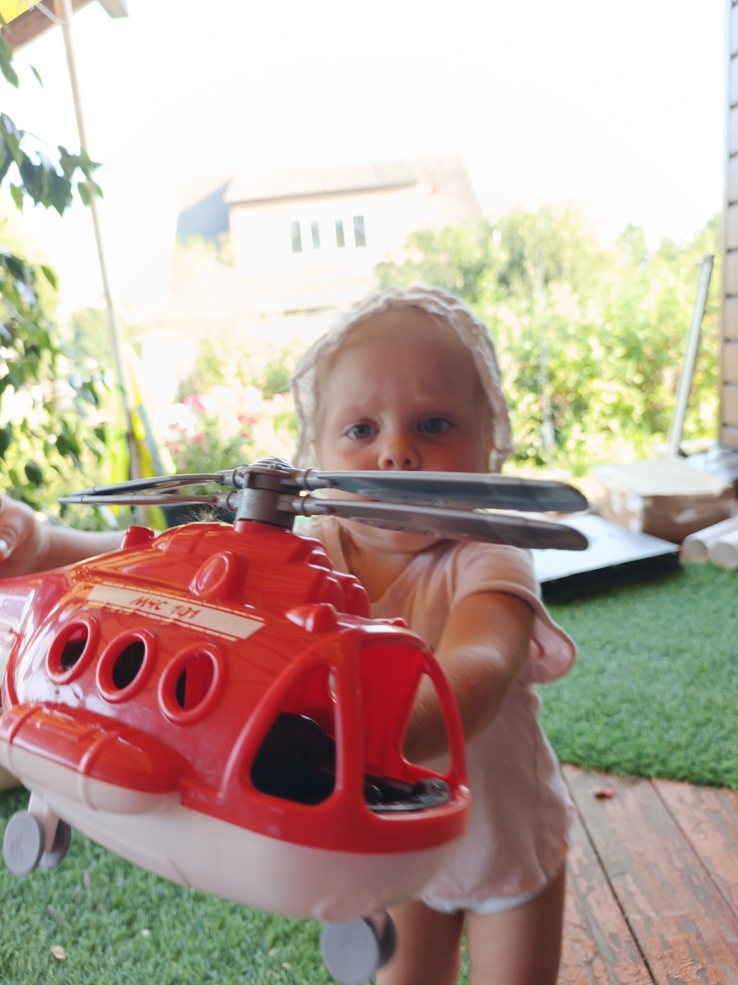 A young child, wearing a white bonnet and a light pink outfit, is holding a red toy helicopter in front of them. The helicopter has a white stripe with "MHC 101" printed on it. The child's face is visible, with blue eyes looking forward. They are standing on a wooden deck covered in green artificial turf. In the background, there are out-of-focus trees and foliage, suggesting an outdoor setting, possibly a yard or garden. A building with windows can be seen in the distance. The lighting indicates it is daytime.