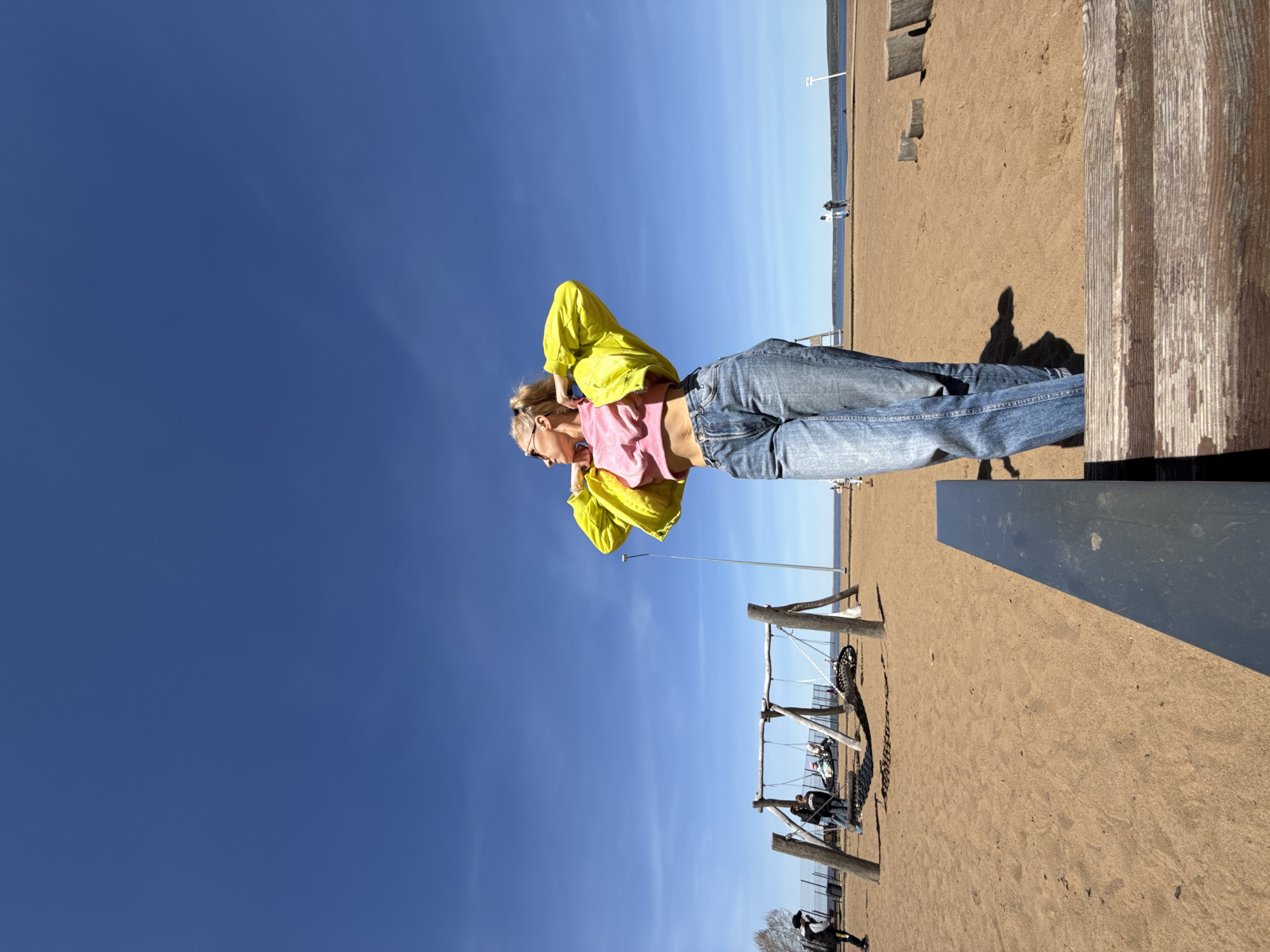 The image depicts a woman standing on a sandy surface with a clear blue sky overhead. She is wearing blue jeans, a pink top, and a bright yellow jacket. Her hair is tied back. She appears to be adjusting the jacket. The location is the beach of the city of Chaikovsky, Russia. In the background, there is a body of water, likely a river or lake, and a sandy beach with a few scattered objects or people. There is a swing set and other infrastructure in the background. A wooden structure is on the right. Shadows suggest a sunny day.