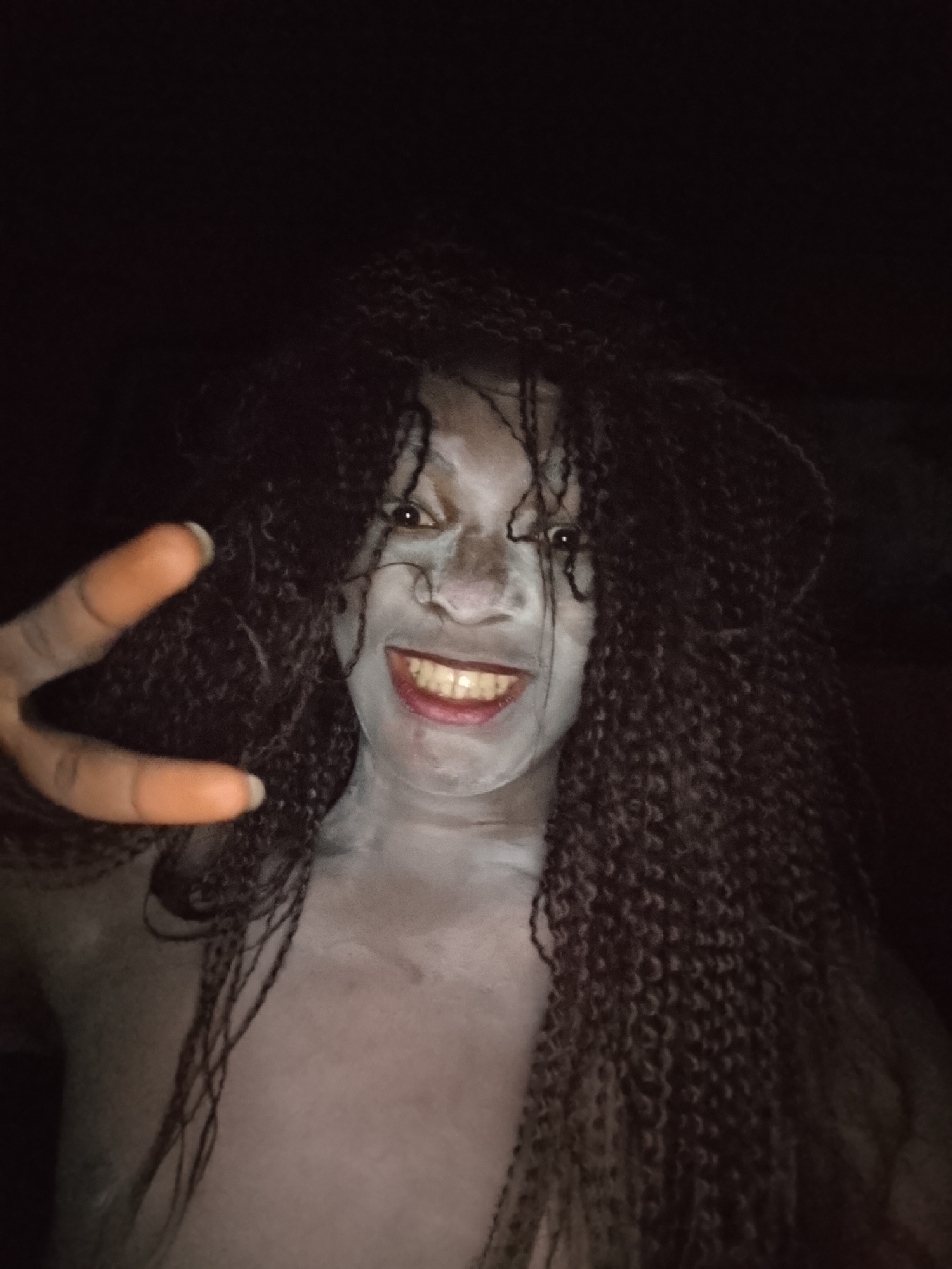 A person with braids, painted white skin, and red lipstick smiles broadly while holding up a peace sign. The background is dark, obscuring details. The location is unknown.