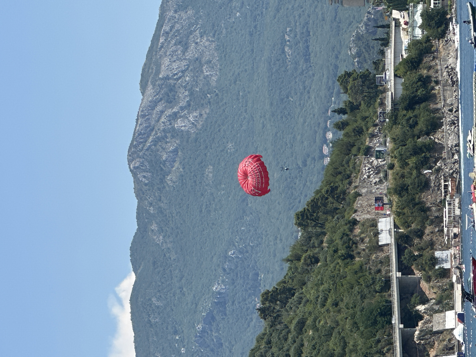 The image shows a red parasail floating in the sky against a backdrop of a lush green mountain and a bright blue sky. A small, dark object, likely a person, is attached to the parasail, and another small dark object, possibly a person or a vehicle, can be seen further in the distance. The parasail appears to be in mid-air, suggesting an activity like parasailing is taking place.
The scene is set outdoors, with the mountain dominating the background. The lower part of the image reveals a coastal area with trees, rocky terrain, and some structures, possibly buildings or cable car stations, running along the hillside. A body of water, likely the sea, is visible on the far right of the image, with several boats docked or anchored. The overall impression is of a sunny day, given the clear blue sky and bright lighting. The location appears to be a scenic, mountainous coastal region, possibly a resort area given the presence of boats and structures. The image is oriented vertically, as if taken from a boat or a lower vantage point looking upwards. There is no visible text in the image.