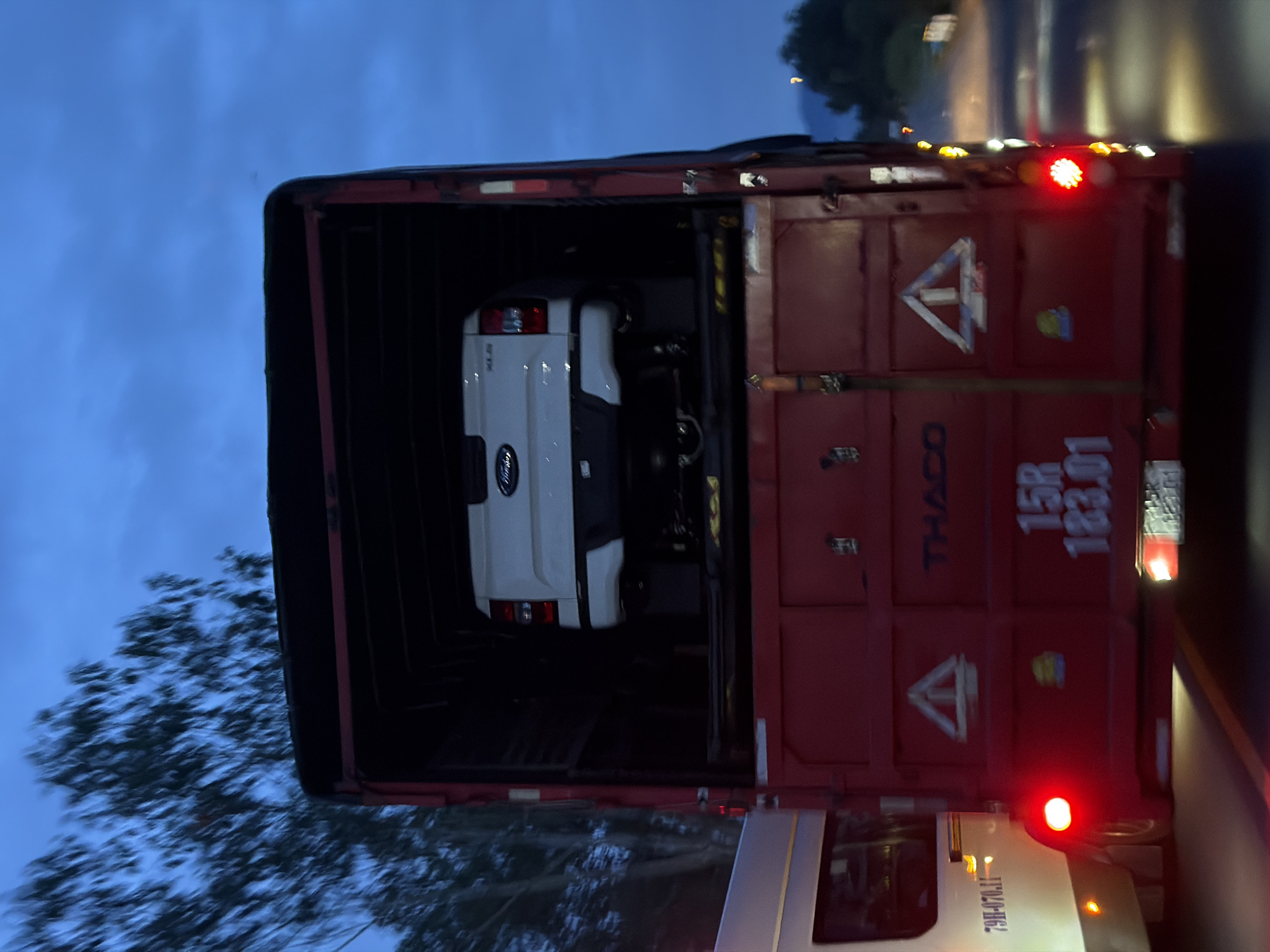A white Ford pickup truck is secured within a red THACO transport container. The container is affixed to a white truck. The photo was taken at dusk in Dien Lac, Vietnam.